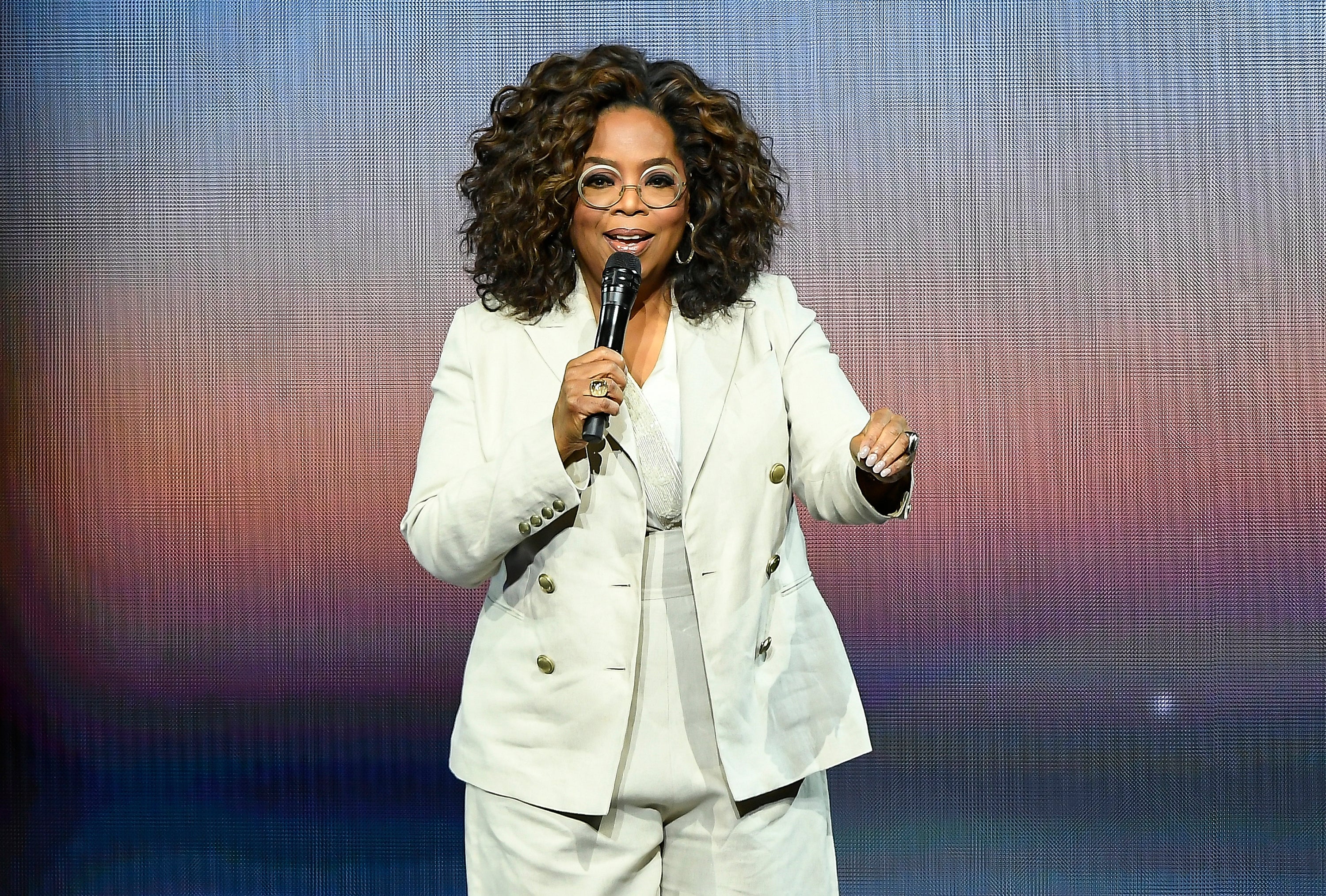A person stands on stage wearing a stylish tailored suit, holding a microphone, and addressing an audience