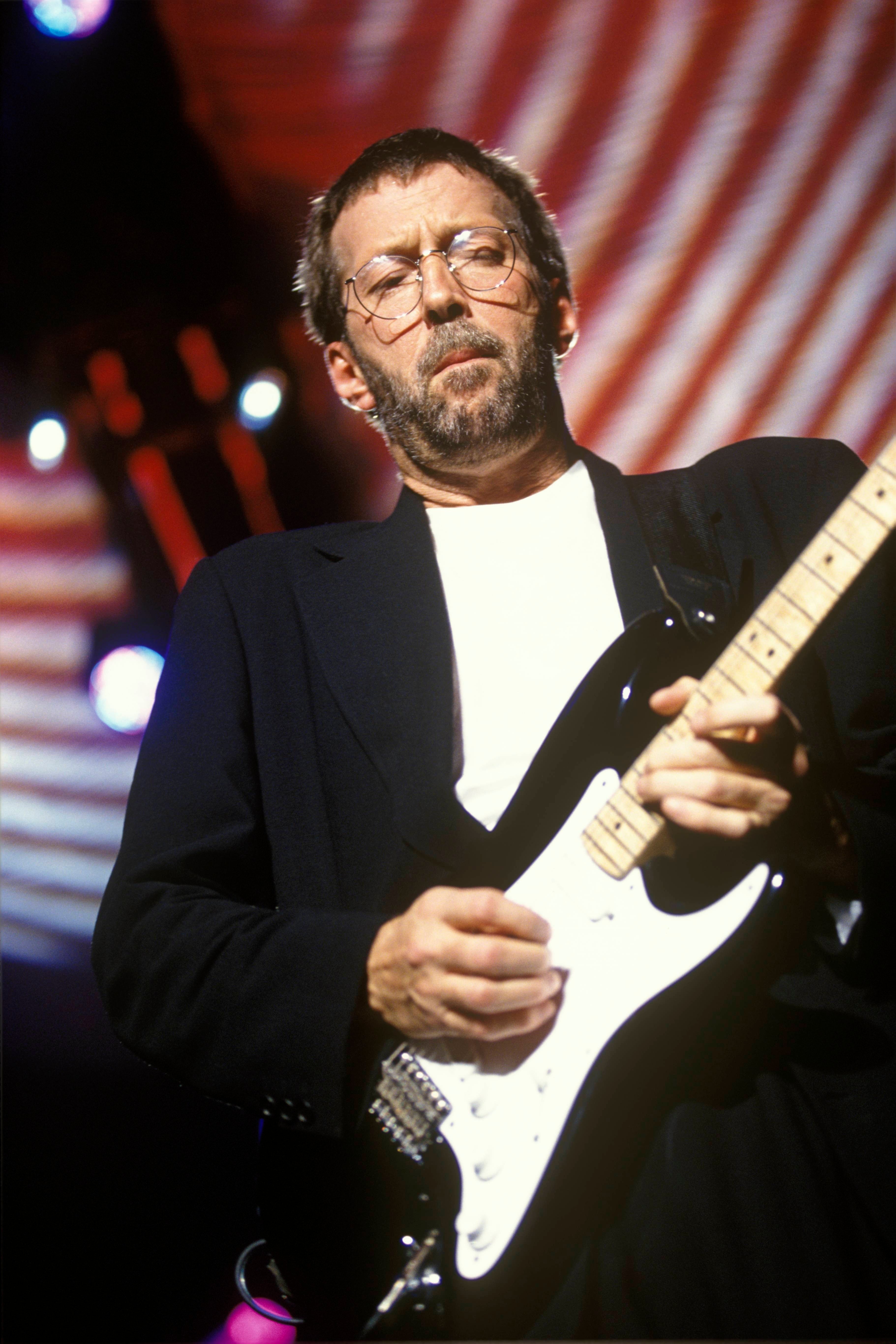 A person in a suit plays an electric guitar on stage, concentrating on the performance, with a dynamic patterned backdrop