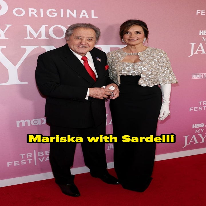 Two people dressed formally on a red carpet. The woman wears a sequined top with white gloves, and the man is in a suit with a patterned tie