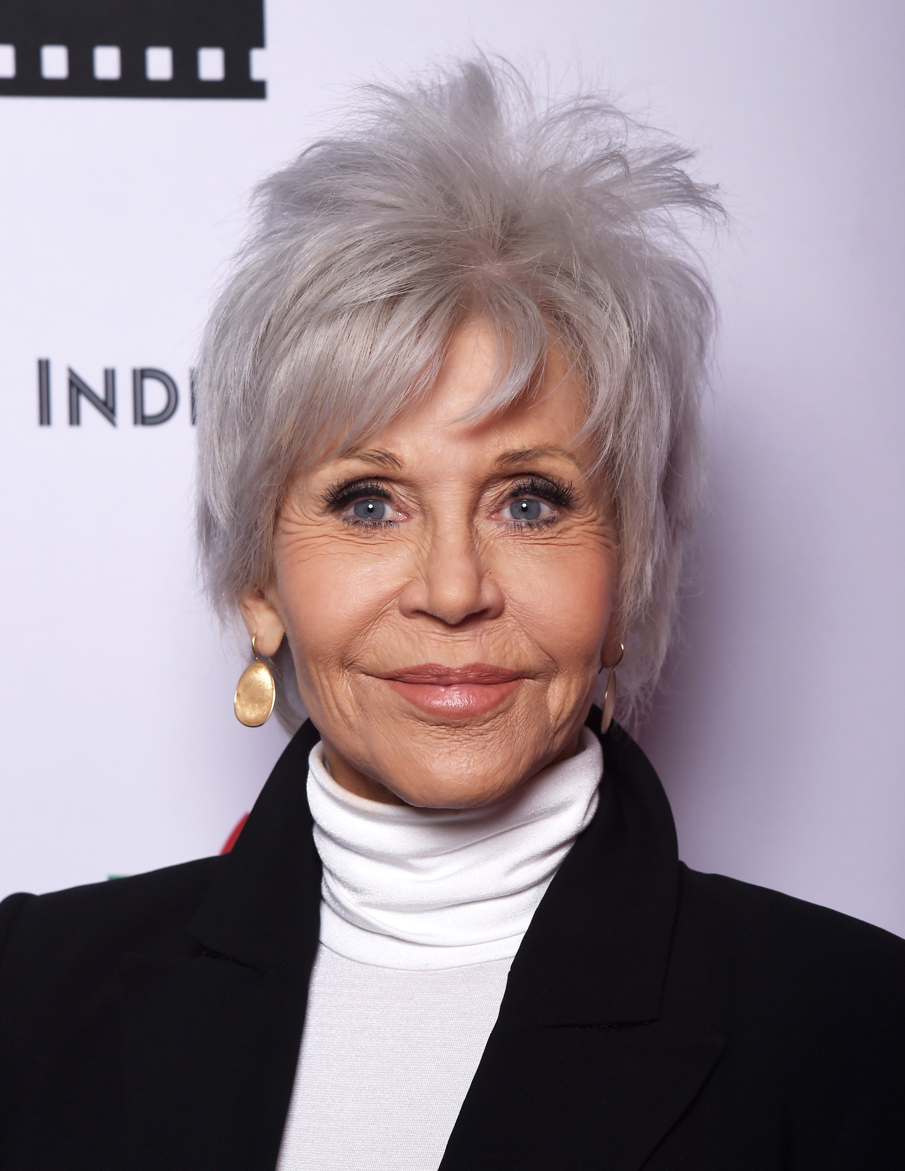 Elderly woman with short, textured hair wears a tailored blazer, white turtleneck, and gold earrings, smiling at a formal event