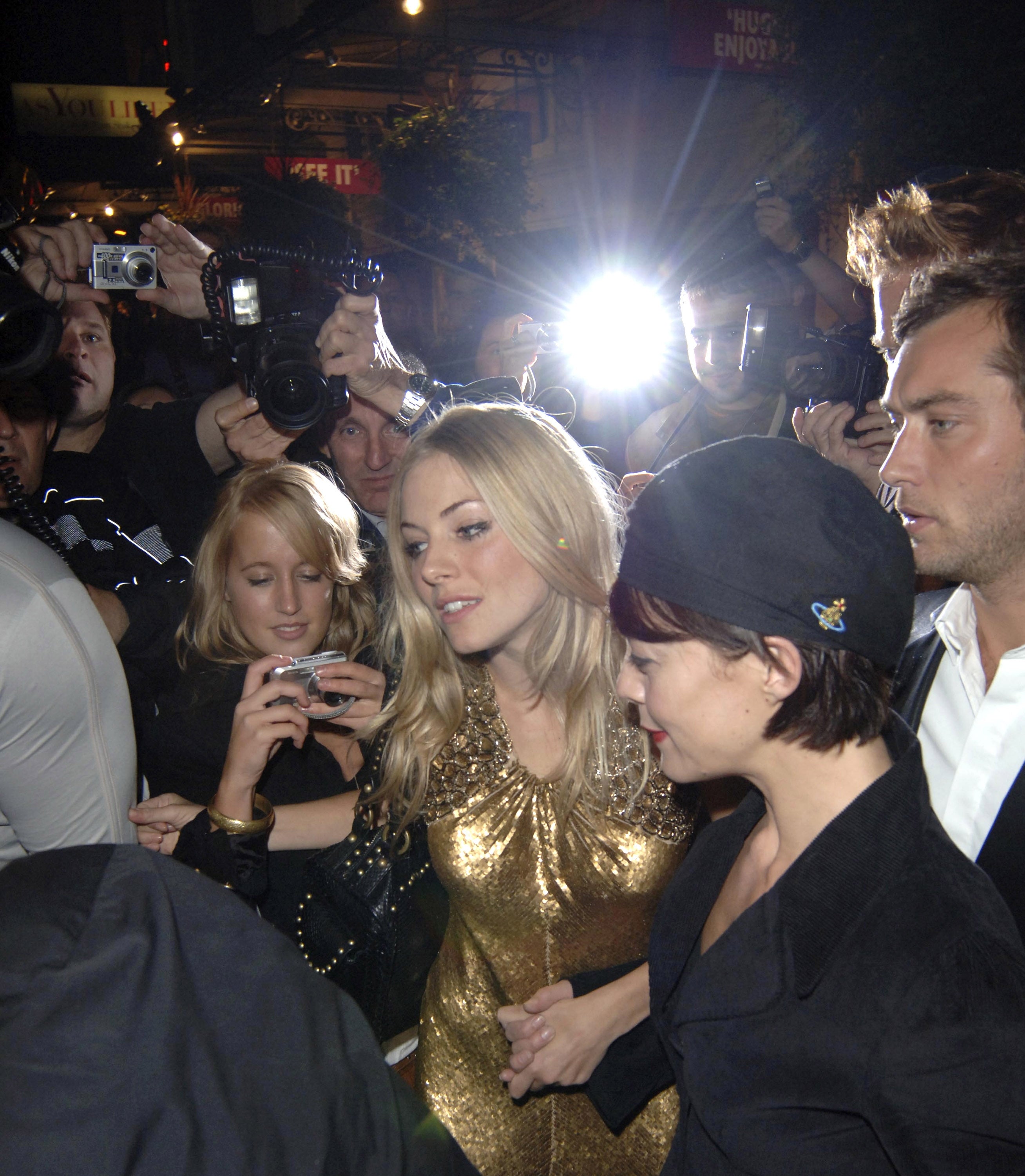 People at an event surrounded by photographers, featuring a woman in a sparkling dress holding hands with another person in a hat
