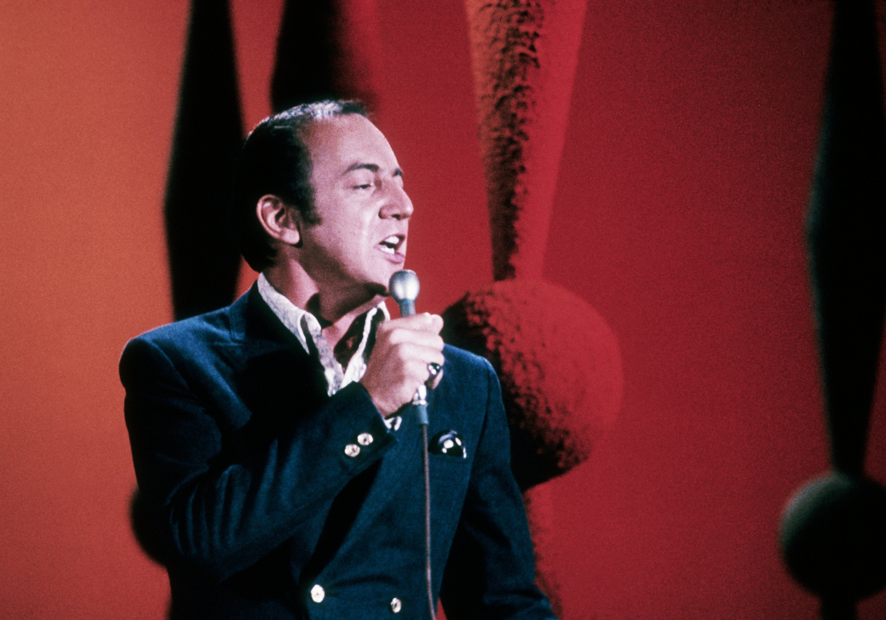 A person sings on stage holding a microphone, wearing a formal jacket. Background features large, abstract decor