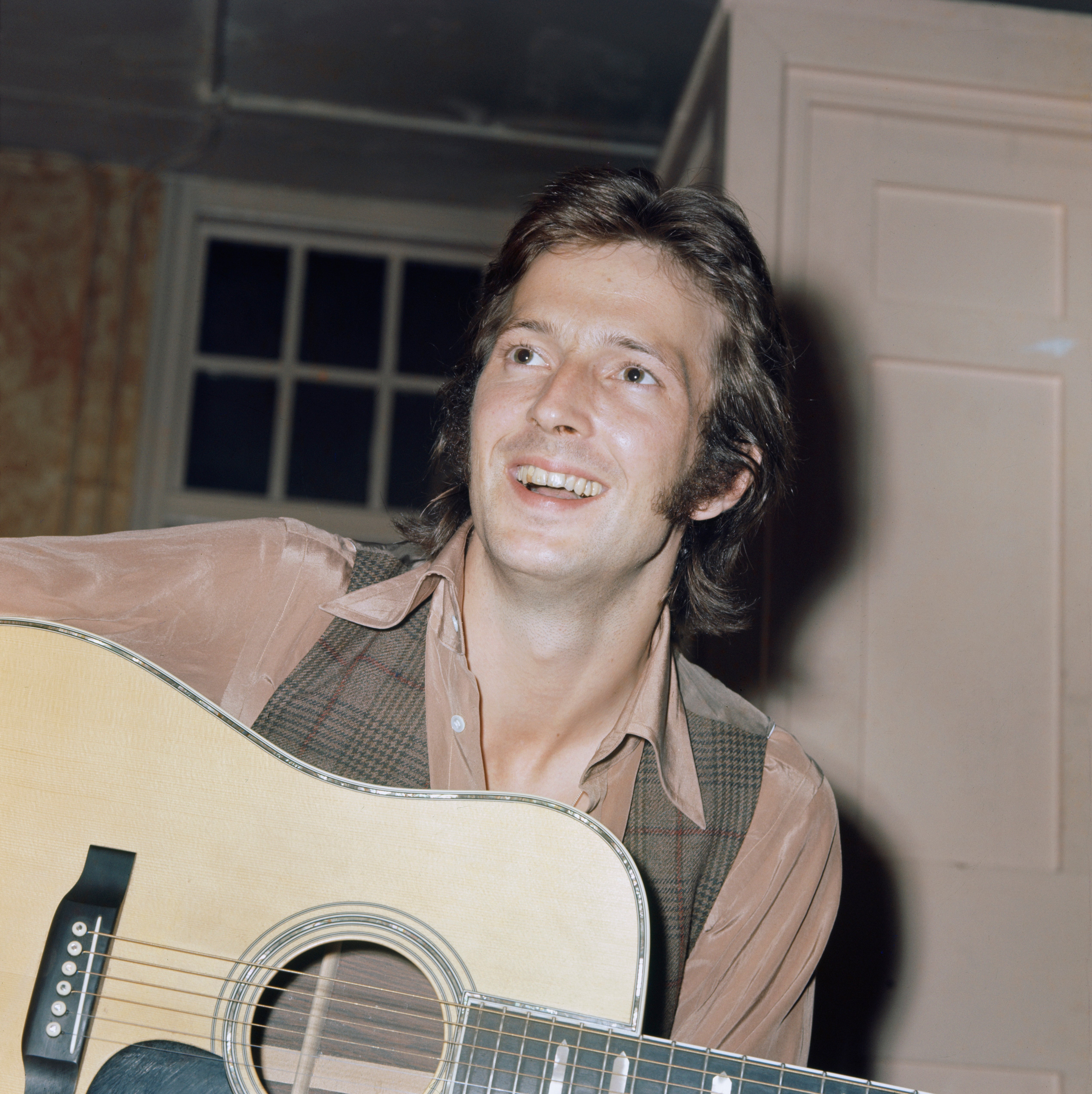 Man smiling while playing an acoustic guitar indoors, wearing a collared shirt and vest