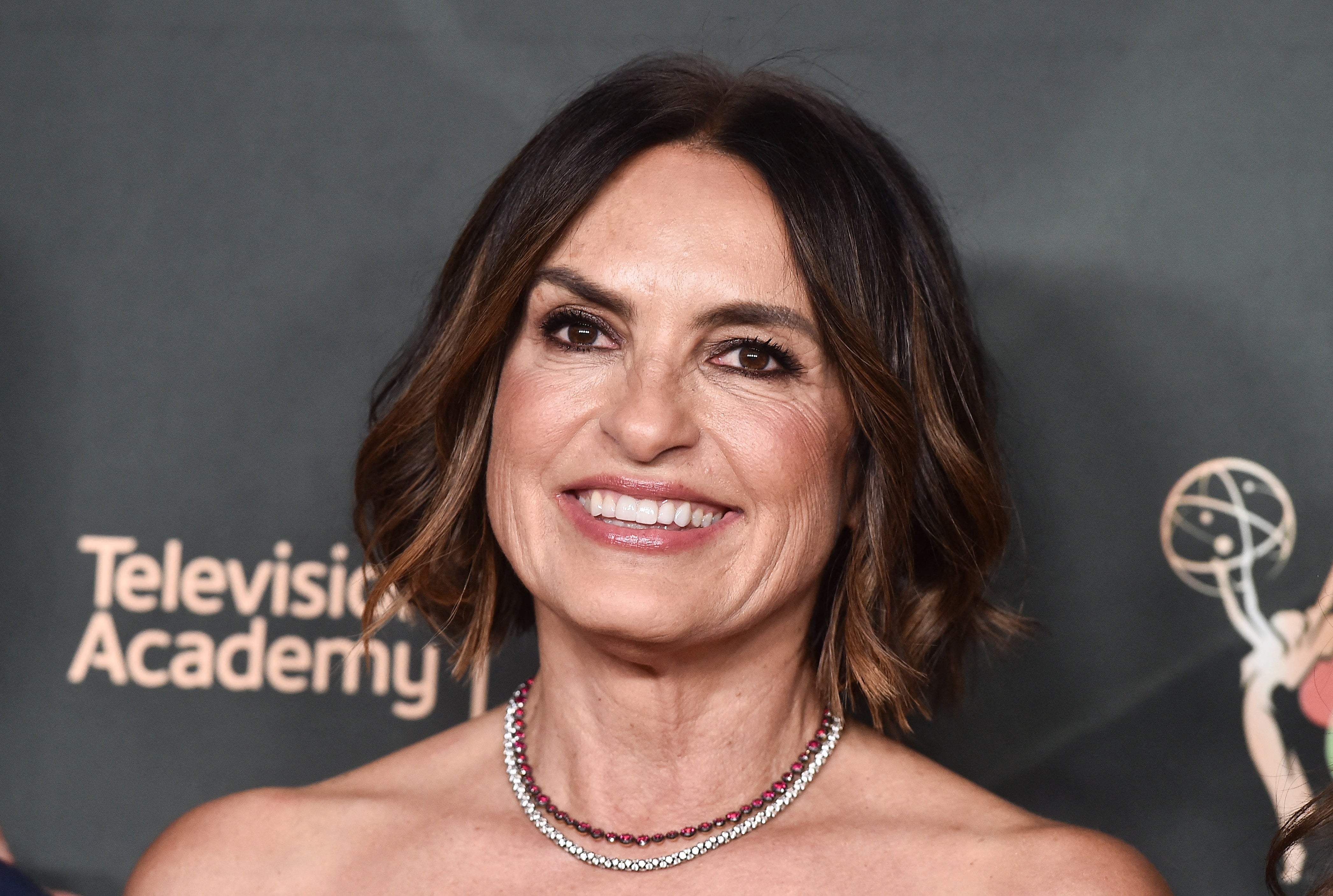 A person smiling on the red carpet in an off-shoulder gown and a beaded necklace, with the Television Academy backdrop in view