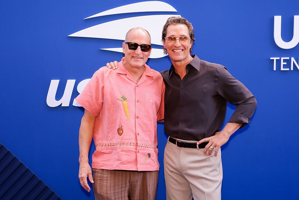 Two people smiling and posing together at an event, both wearing sunglasses. One wears a casual embroidered shirt and the other a sleek, button-down shirt