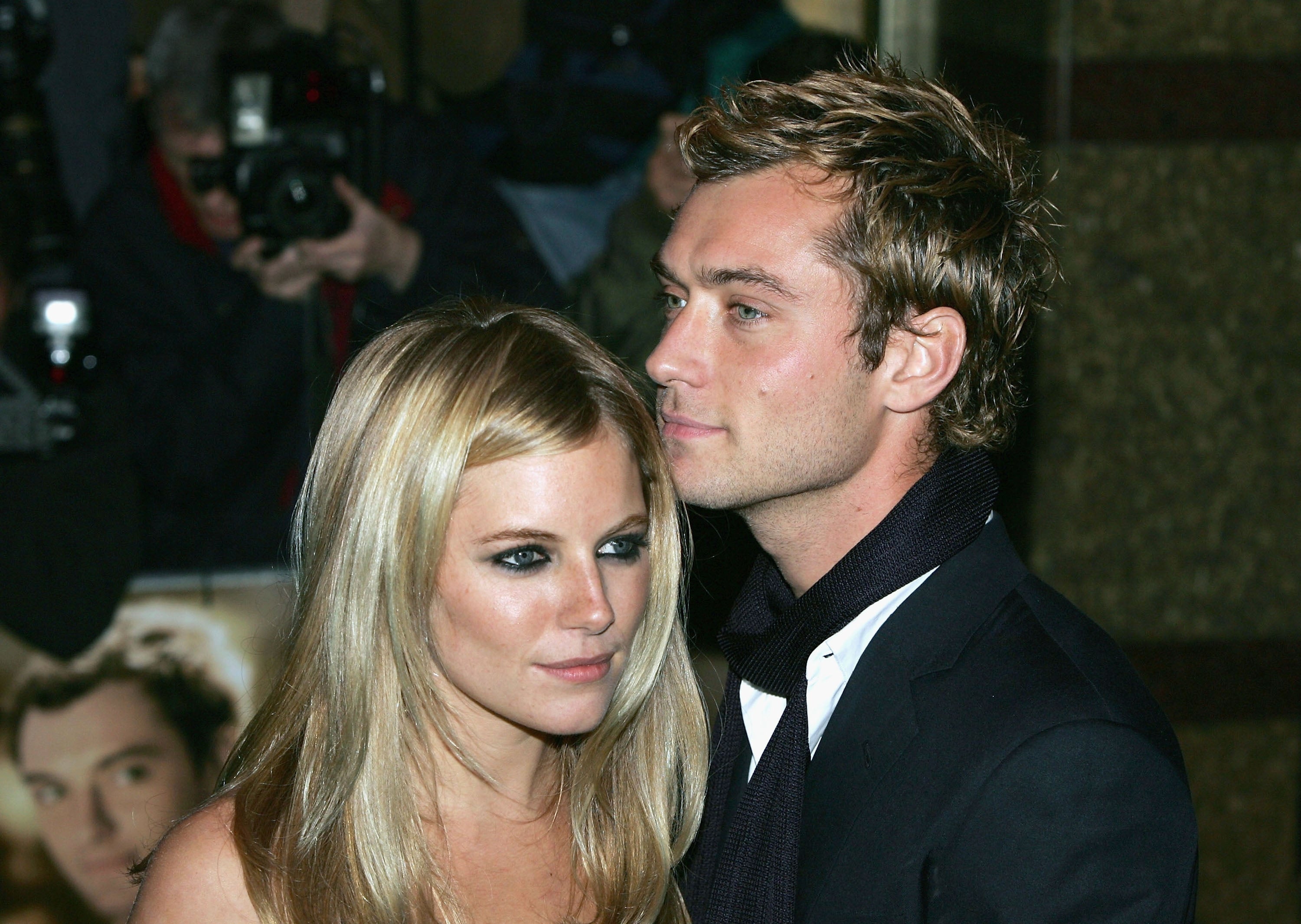 A couple poses at an event. The man wears a dark suit with a scarf, and the woman has long hair and wears a stylish outfit. Photographers in the background