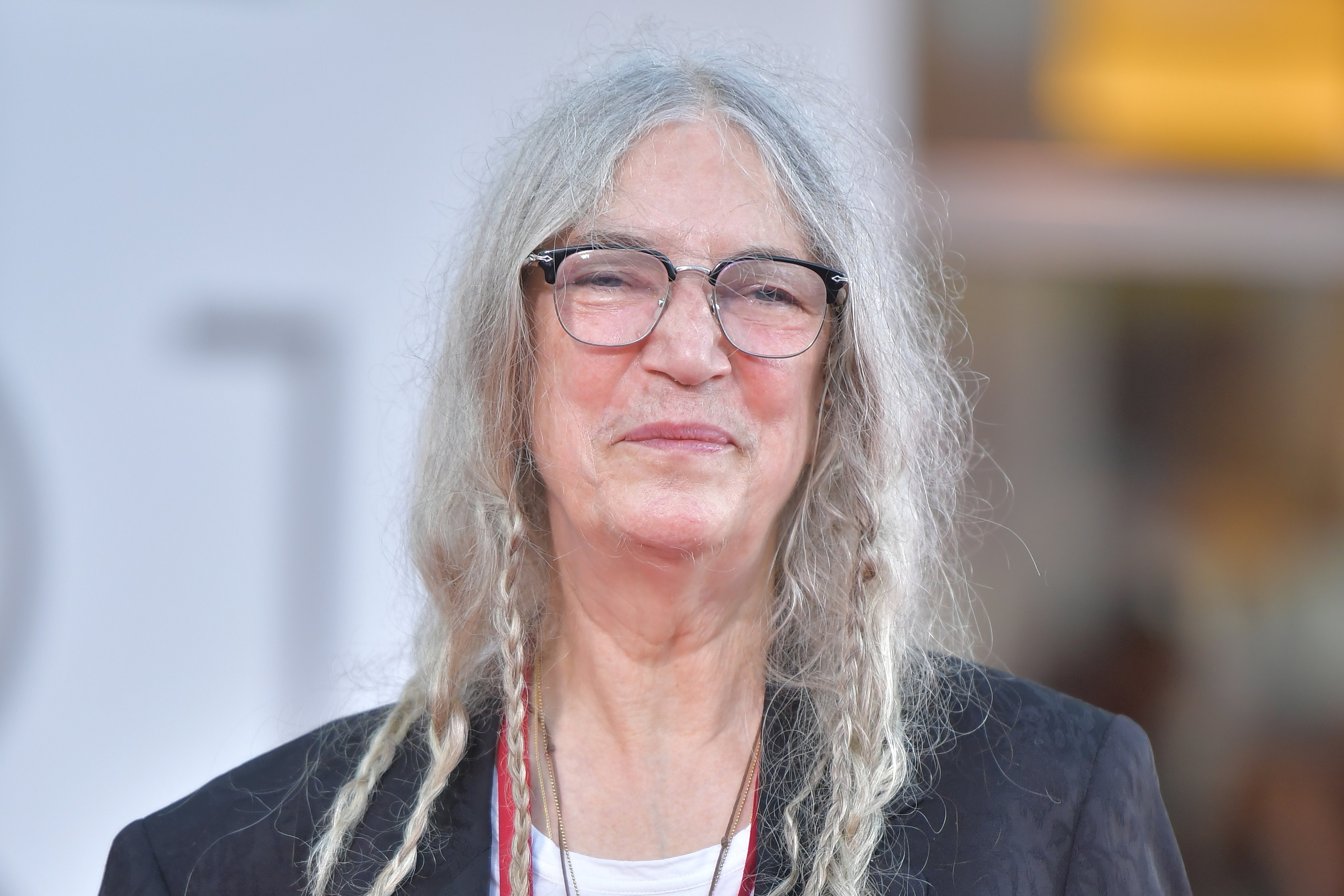 Person with long hair, glasses, and casual attire smiles on a red carpet