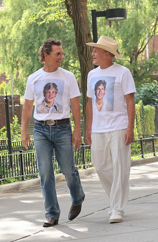 Two people walking and smiling, wearing t-shirts featuring childhood photos. One wears jeans, the other has light striped pants and a hat