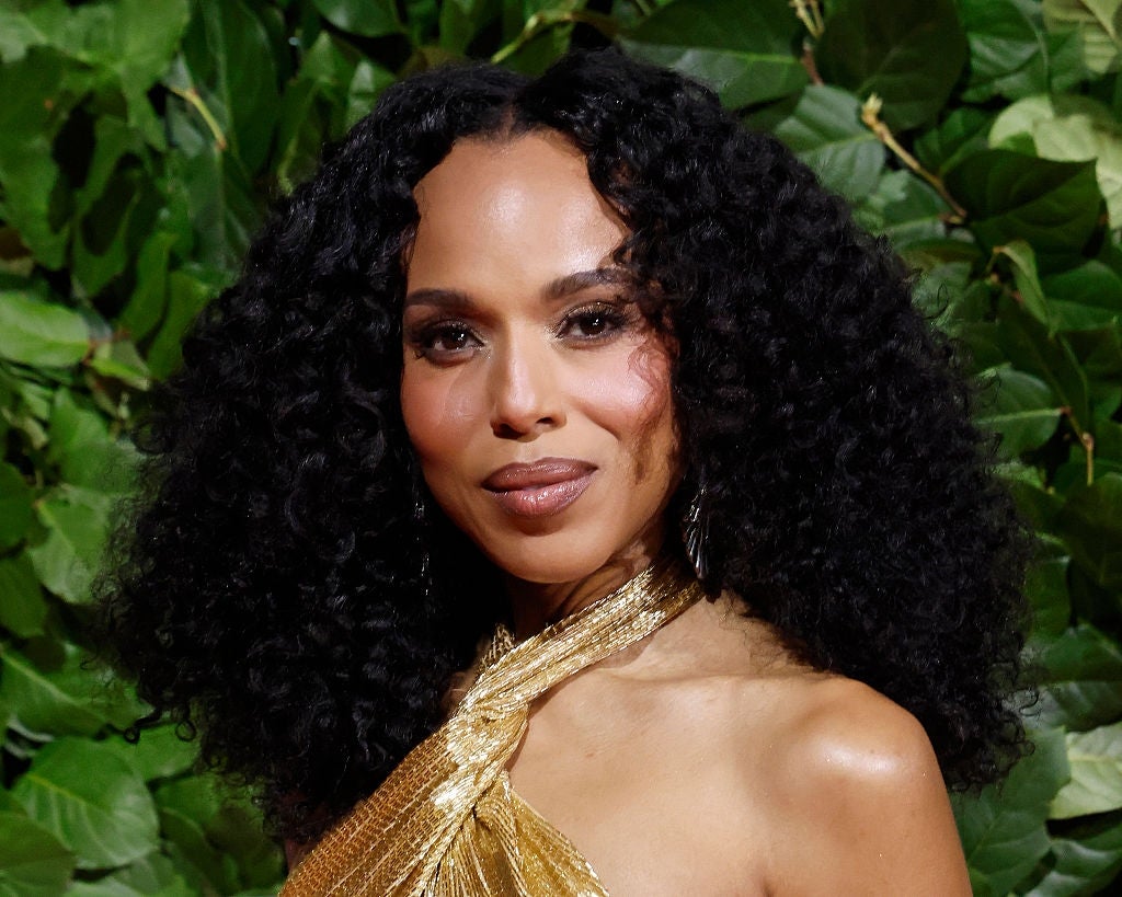 A woman with curly hair wears a gold, one-shoulder gown, posing in front of a leafy backdrop