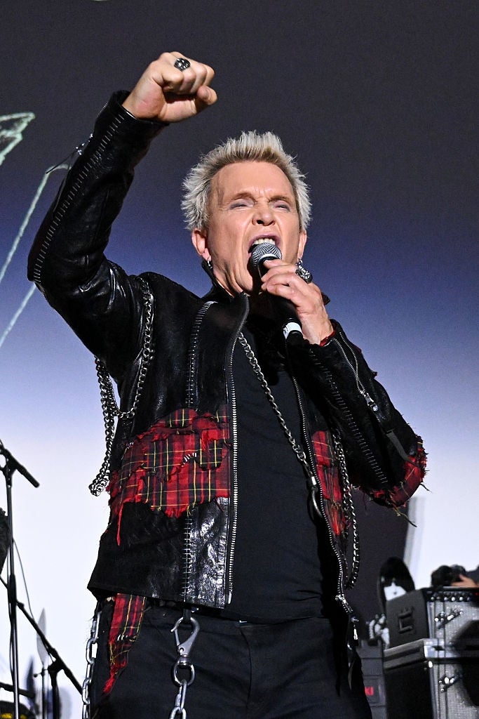 Person passionately performing on stage, wearing a black and red patterned jacket with chains, holding a microphone and raising a fist