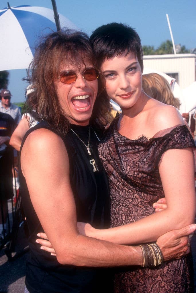 Two people smiling at an outdoor event, one wearing a sleeveless top and sunglasses, the other in an off-shoulder lace dress with short hair