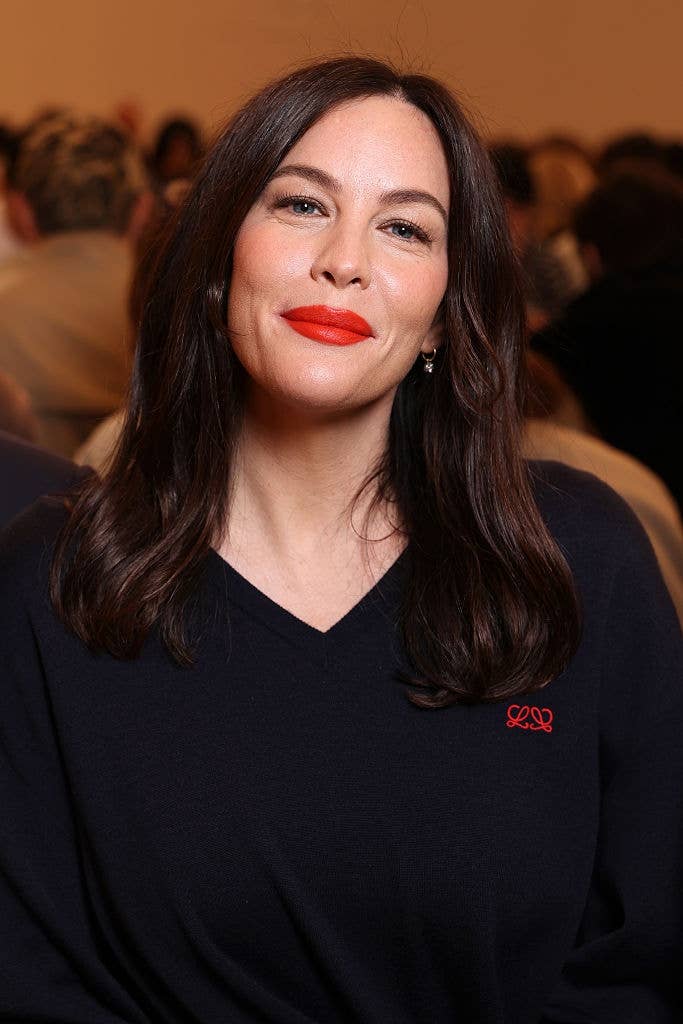 Person with long, dark hair wearing a dark V-neck sweater, seated in a crowded indoor setting, smiling with bright lipstick
