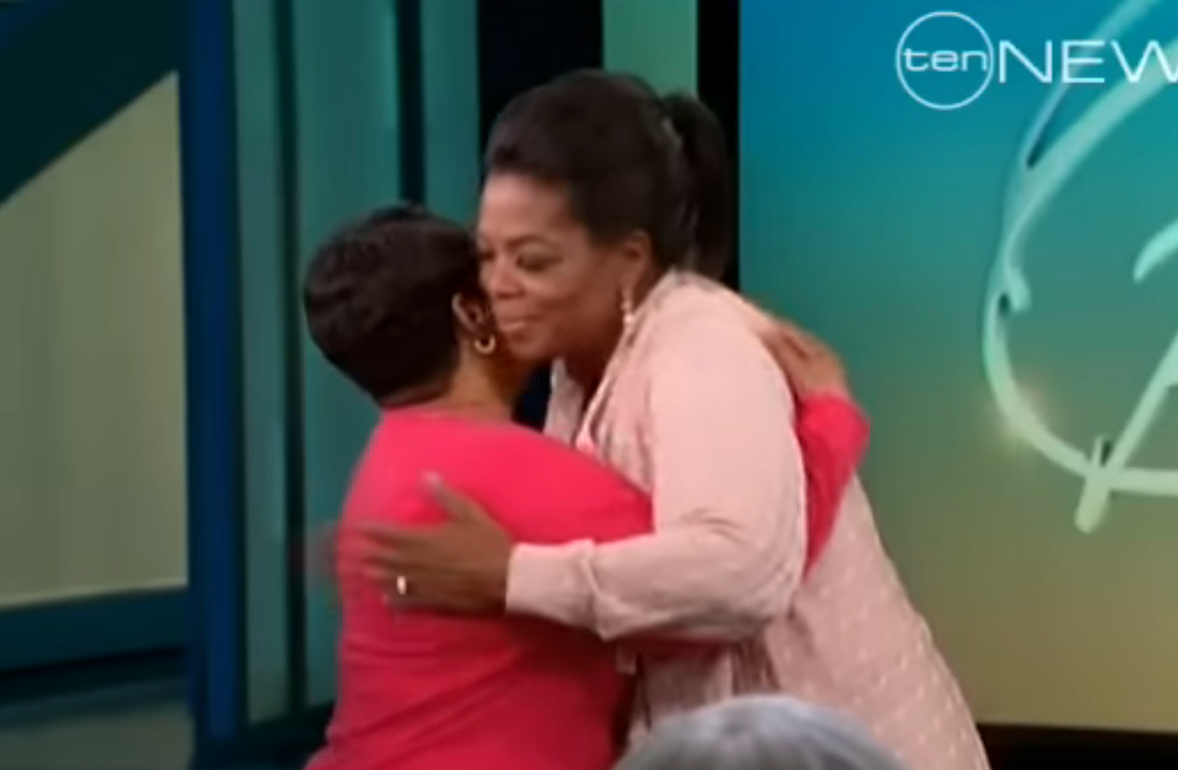 Two women embrace warmly on a talk show set, one in a patterned long dress