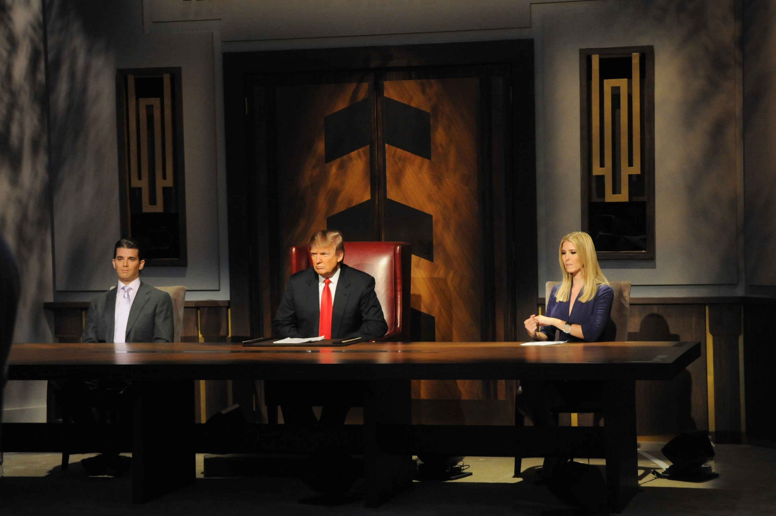 Three people sit at a long table in a formal setting, reminiscent of a boardroom scene