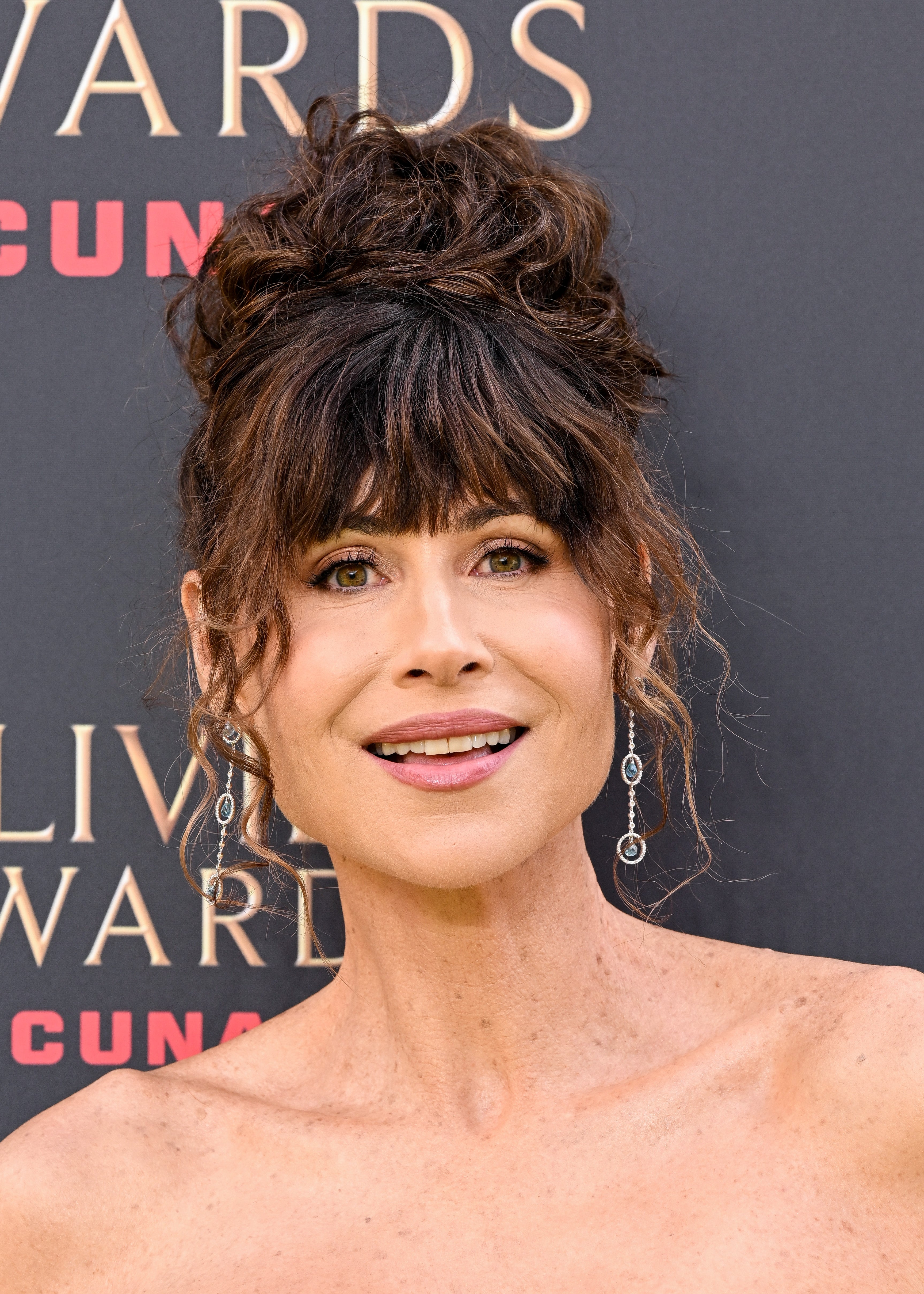 A person with curly updo hairstyle and dangling earrings at an award event, smiling and wearing an off-the-shoulder dress