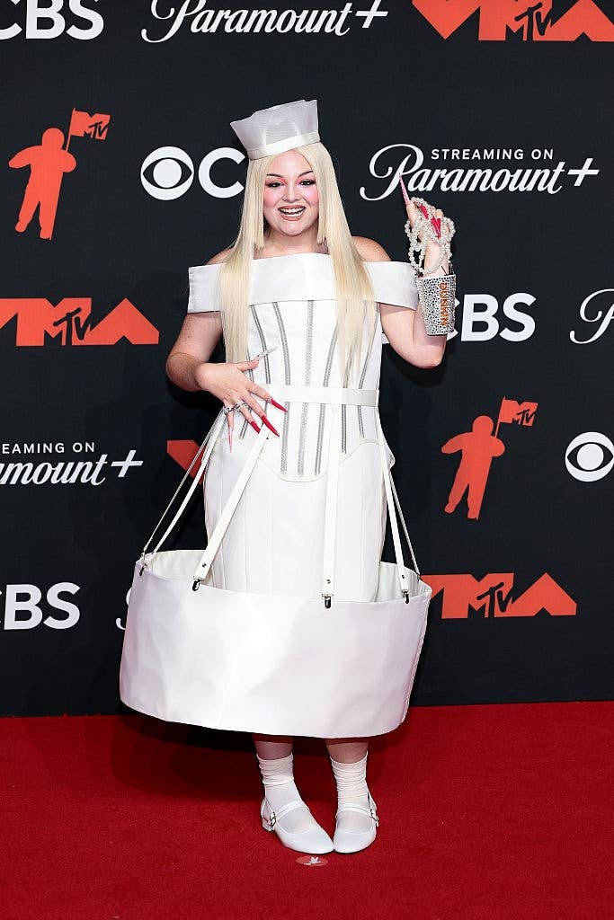 Meg on red carpet in an avant-garde white outfit resembling a coffee cup , holding a silver Dunkin cup clutch, and wearing long blonde hair