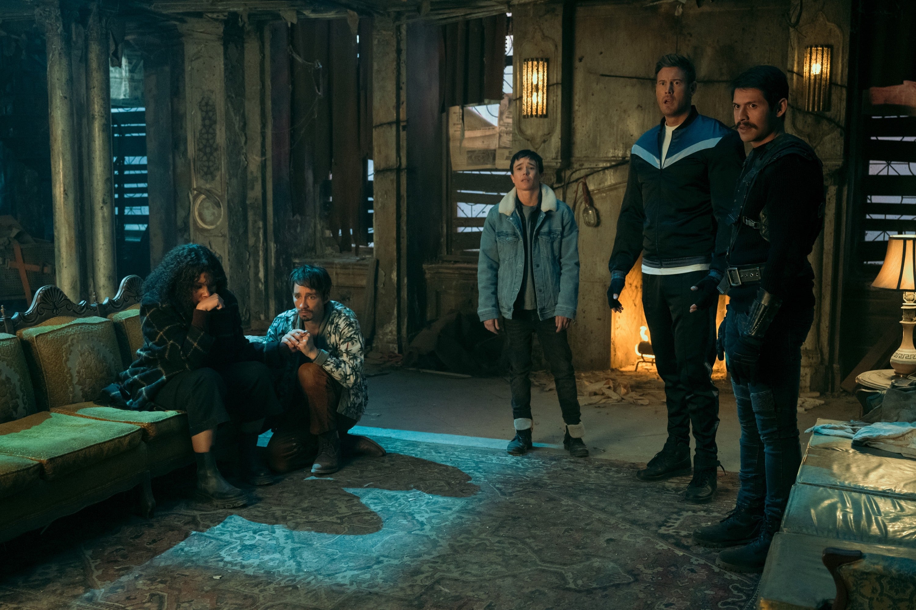 Scene from a TV show with five characters in a dimly-lit, worn room, wearing casual and rugged outfits, suggesting a dramatic moment