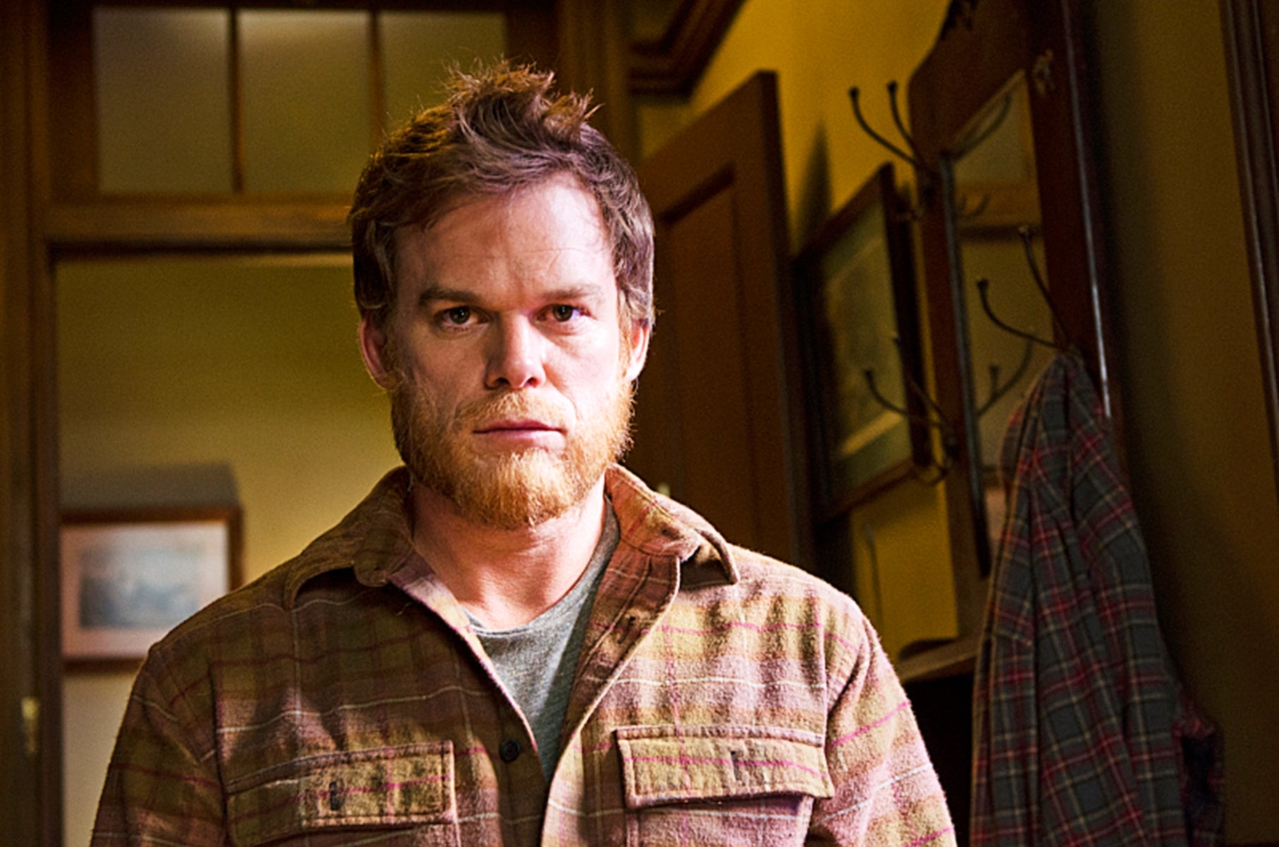 A man with a rugged appearance stands indoors in a casual plaid shirt, appearing contemplative