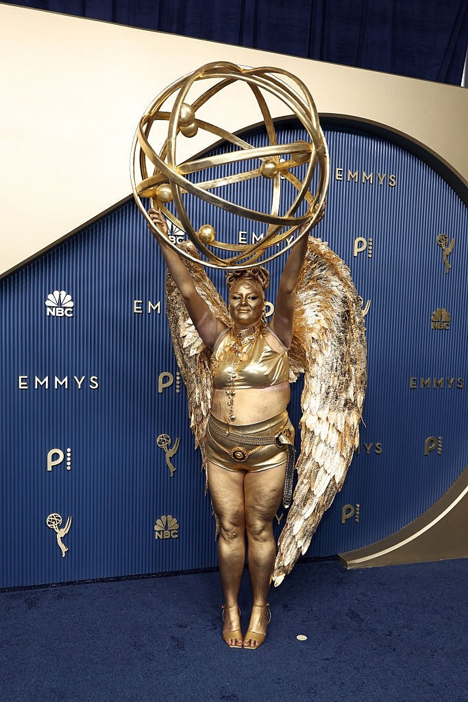 Meg in gold outfit and wings poses with arms raised, holding a spherical structure