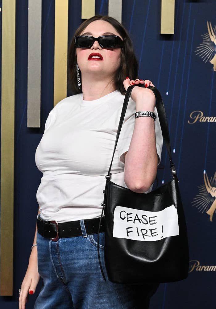 Meg with sunglasses poses confidently on red carpet, wearing a white t-shirt and jeans, holding a bag with "CEASE FIRE!" sign
