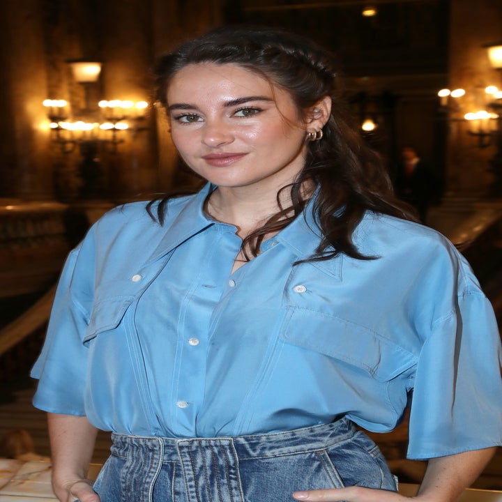 Person wearing a blue shirt and denim skirt stands in an elegant indoor setting, smiling slightly with hands in pockets