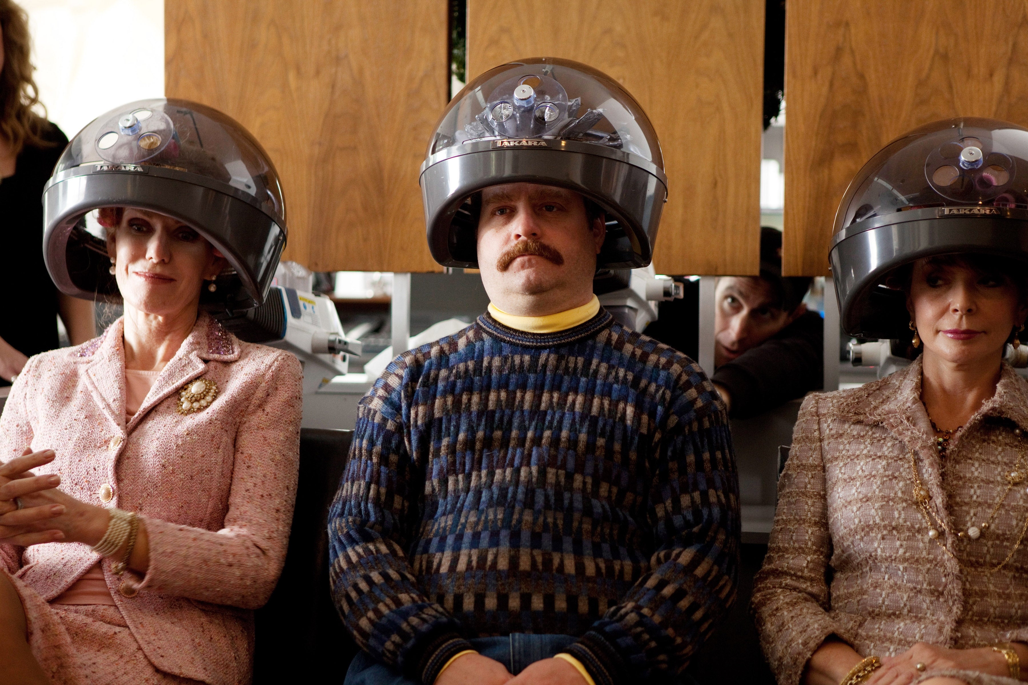 Three people under hair dryers, with one person wearing a patterned sweater and mustache, sitting between two women in elegant suits