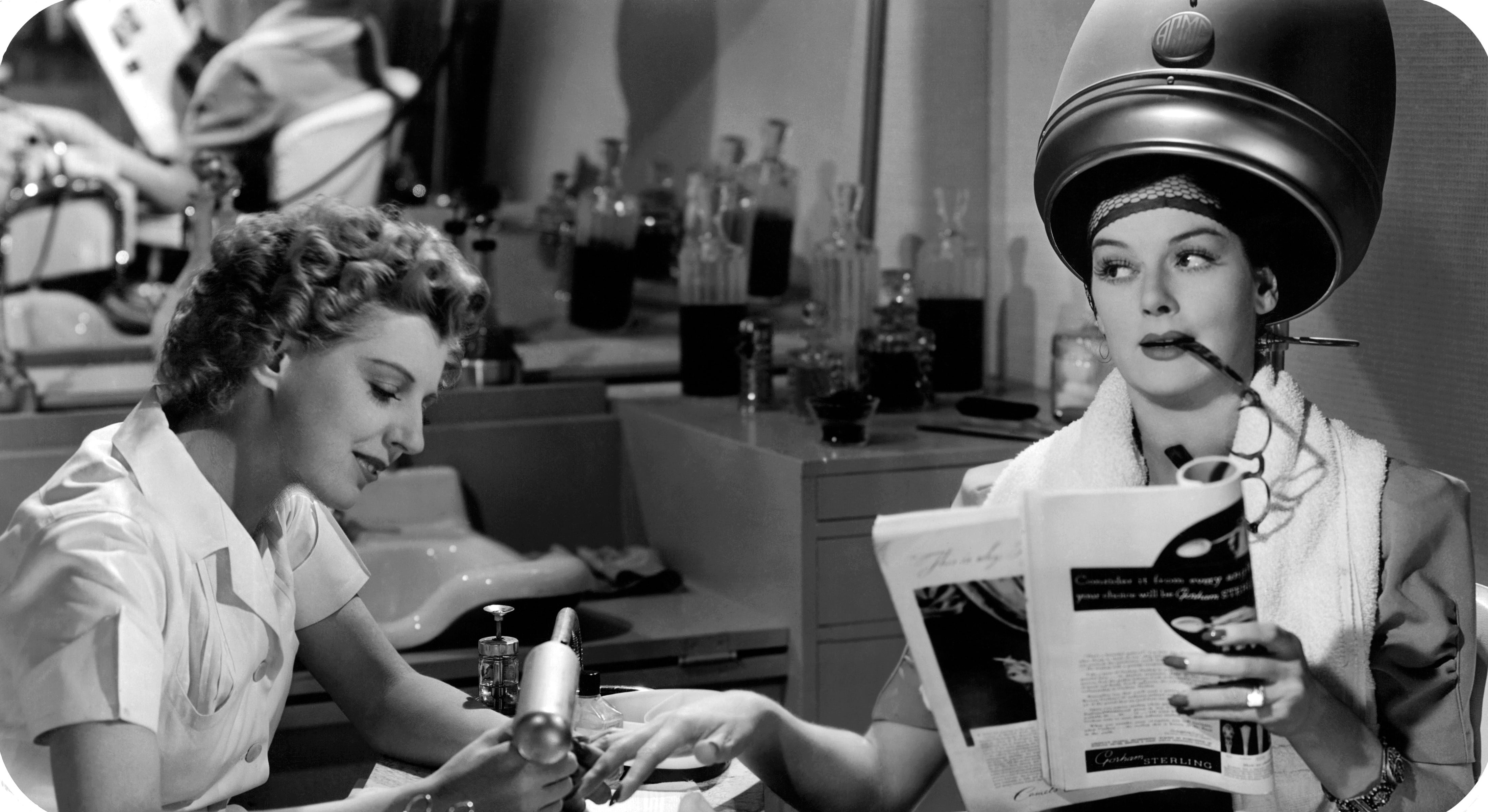 A vintage beauty salon scene with a woman under a hairdryer reading, while a manicurist works on her nails