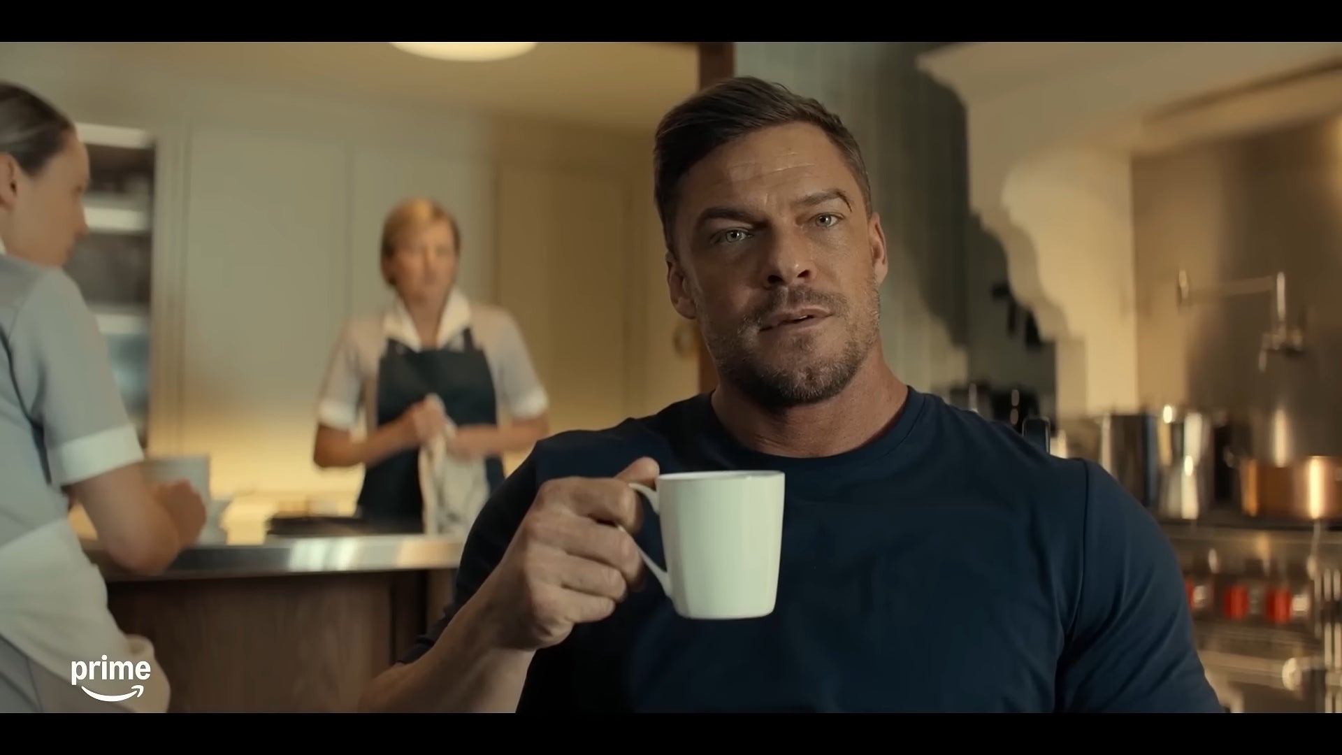 A antheral   with a muscular physique  sits successful  a kitchen, holding a mug. Two radical   are blurred successful  the background, 1  wearing an apron