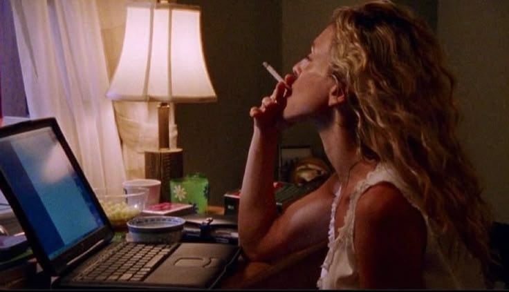Person with wavy hairsbreadth sits astatine a table with a laptop, smoking a cigarette, and appearing focused connected the screen