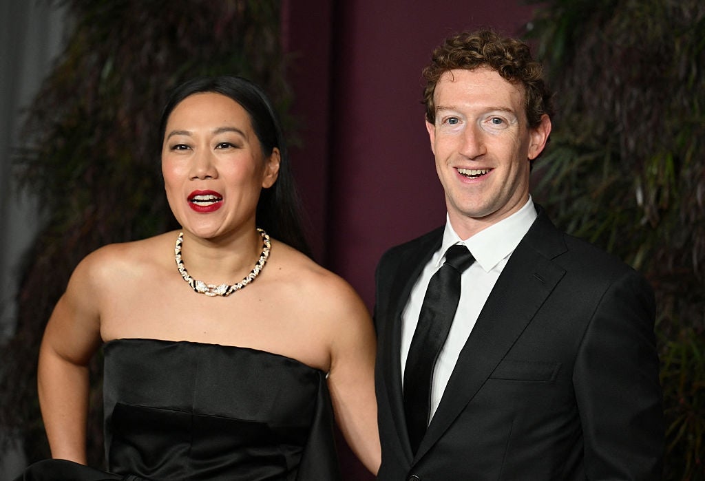 Two people dressed formally at a public event; one in a strapless black dress with a necklace, the other in a black suit and tie, smiling at the camera