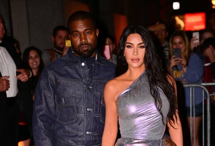 A couple poses on the red carpet. The woman wears a one-shoulder shiny dress, and the man wears a denim jacket