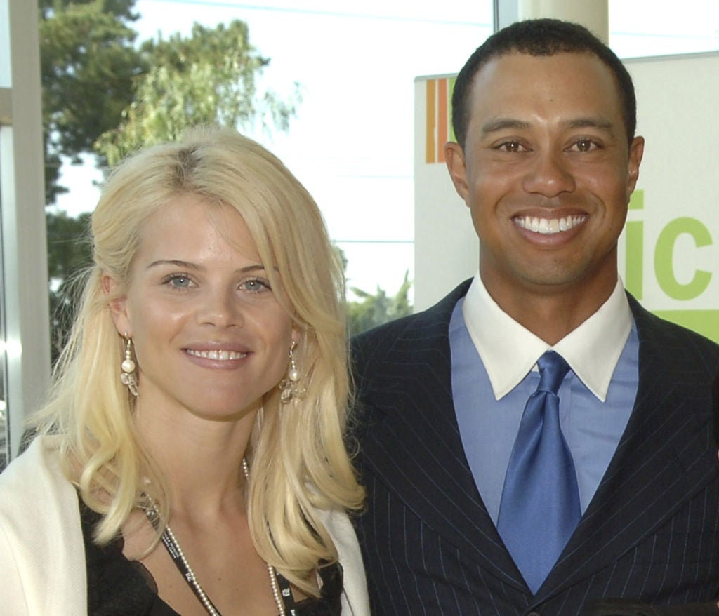 Two individuals at an event, one wearing a striped suit with a blue tie, and the other in a white top with layered necklaces and long earrings