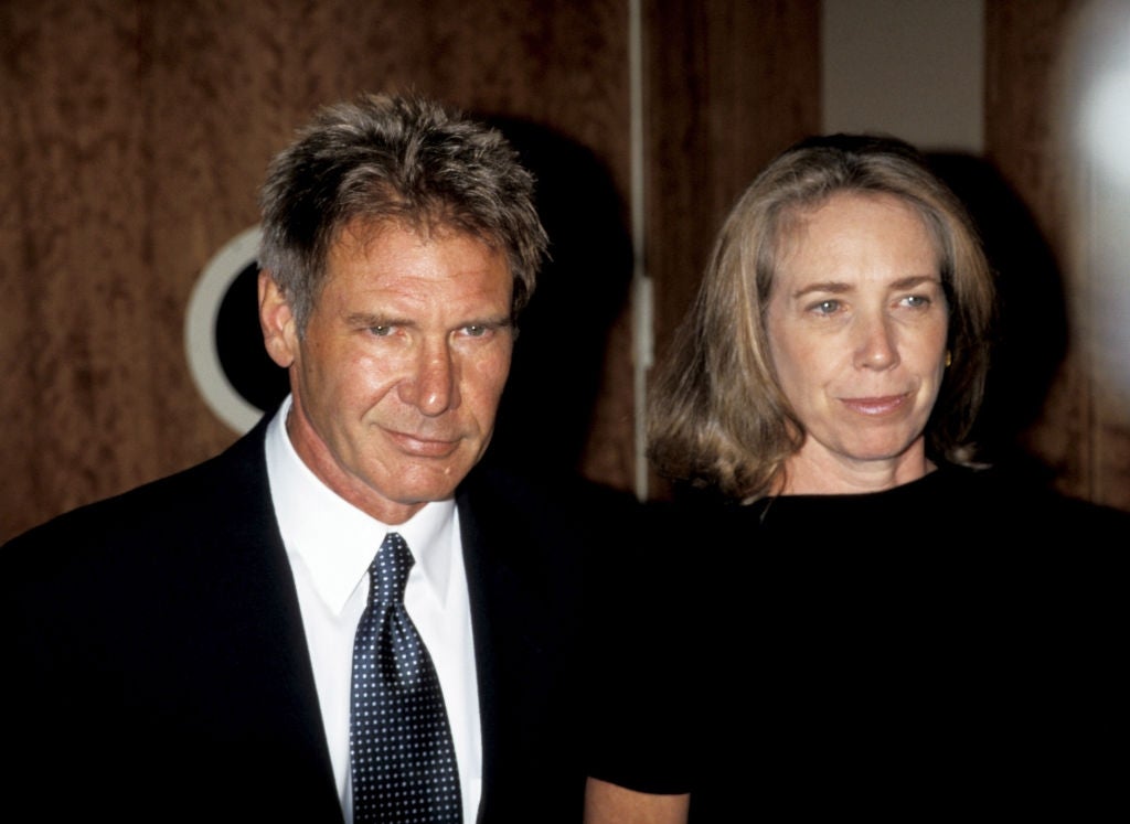 A man and woman stand together, both wearing formal attire, with the man in a suit and tie. They are posing at a formal event or gathering