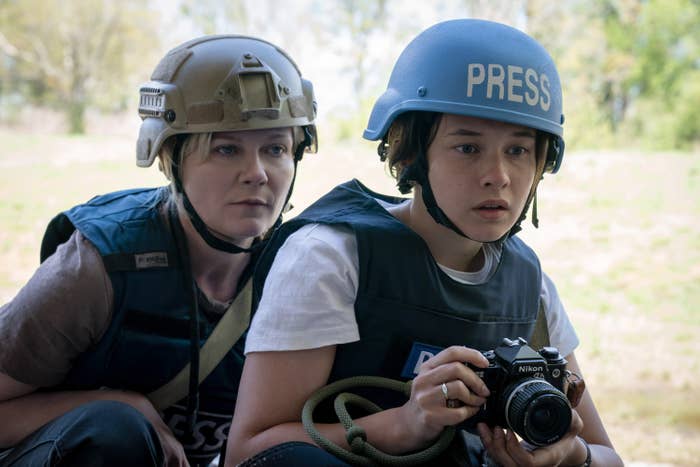 Two journalists wearing helmets and press vests crouch outdoors. One holds a camera, appearing focused on a scene