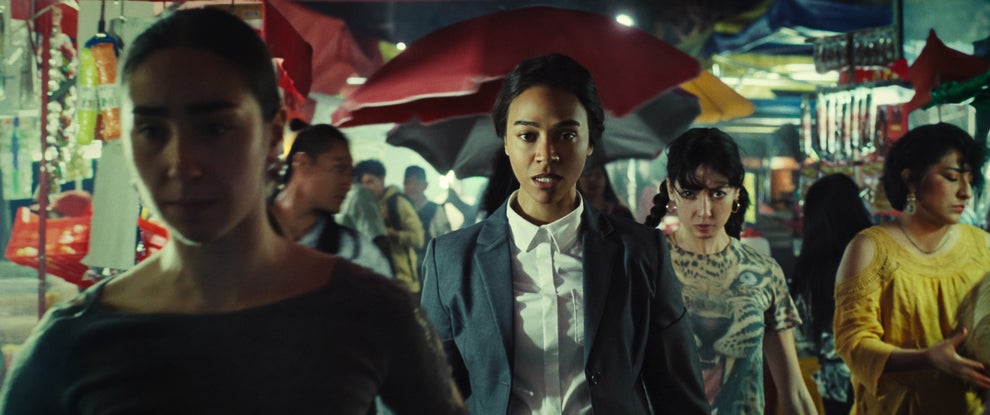 A woman in a business suit walks confidently through a busy market, surrounded by people carrying umbrellas