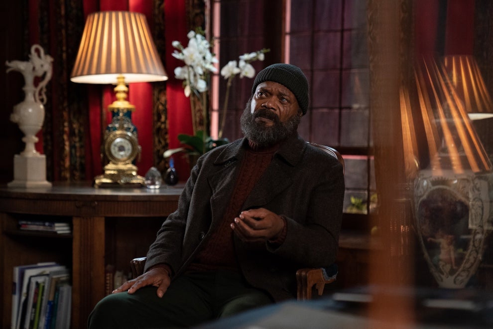 Person in warm, layered clothing and beanie, seated in a room with ornate decor, discussing or gesturing