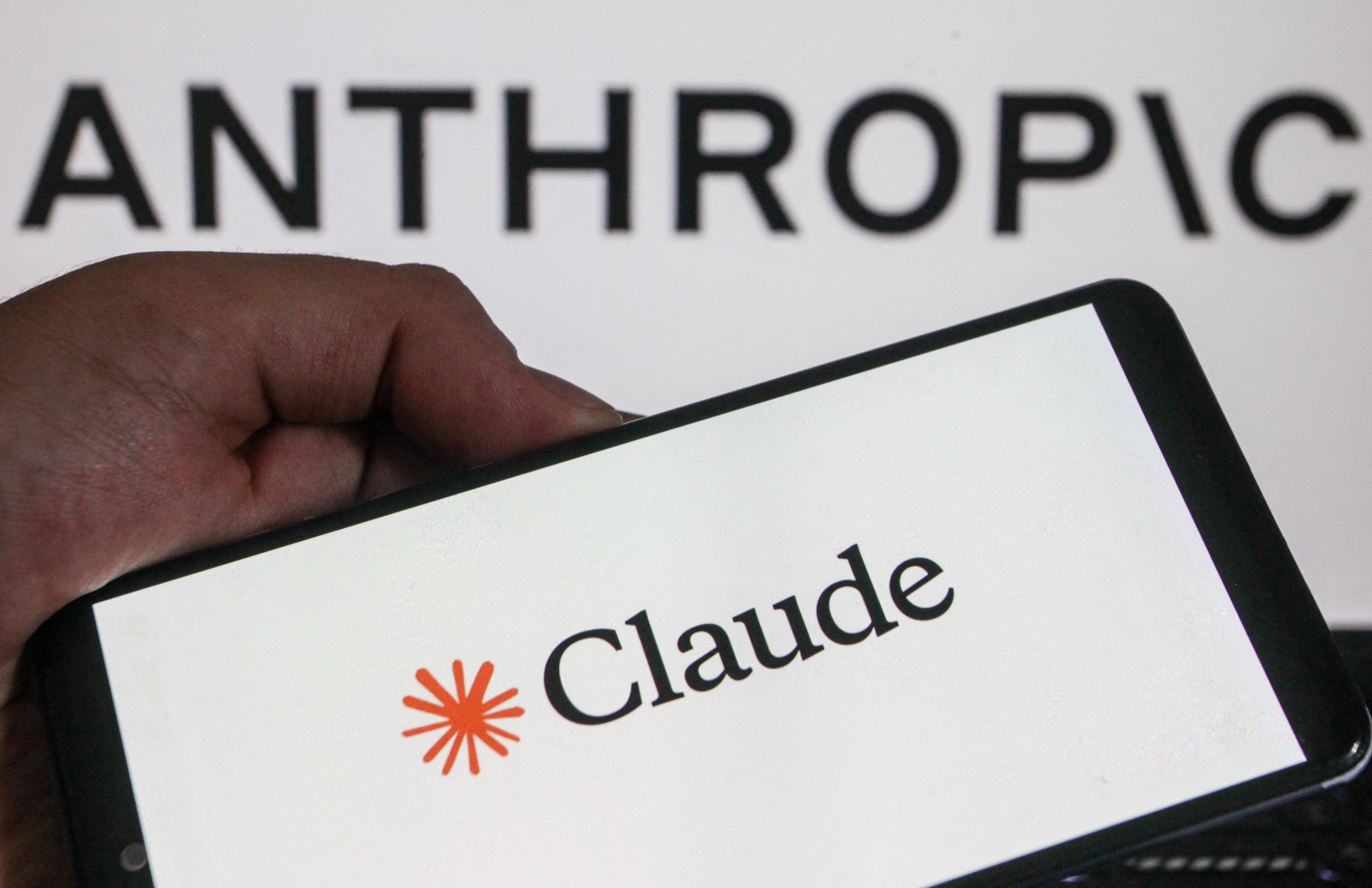 Hånd som holder en smarttelefon som viser "Claude" med en oransje stjernelignende logo. Bakgrunnen inneholder ordet