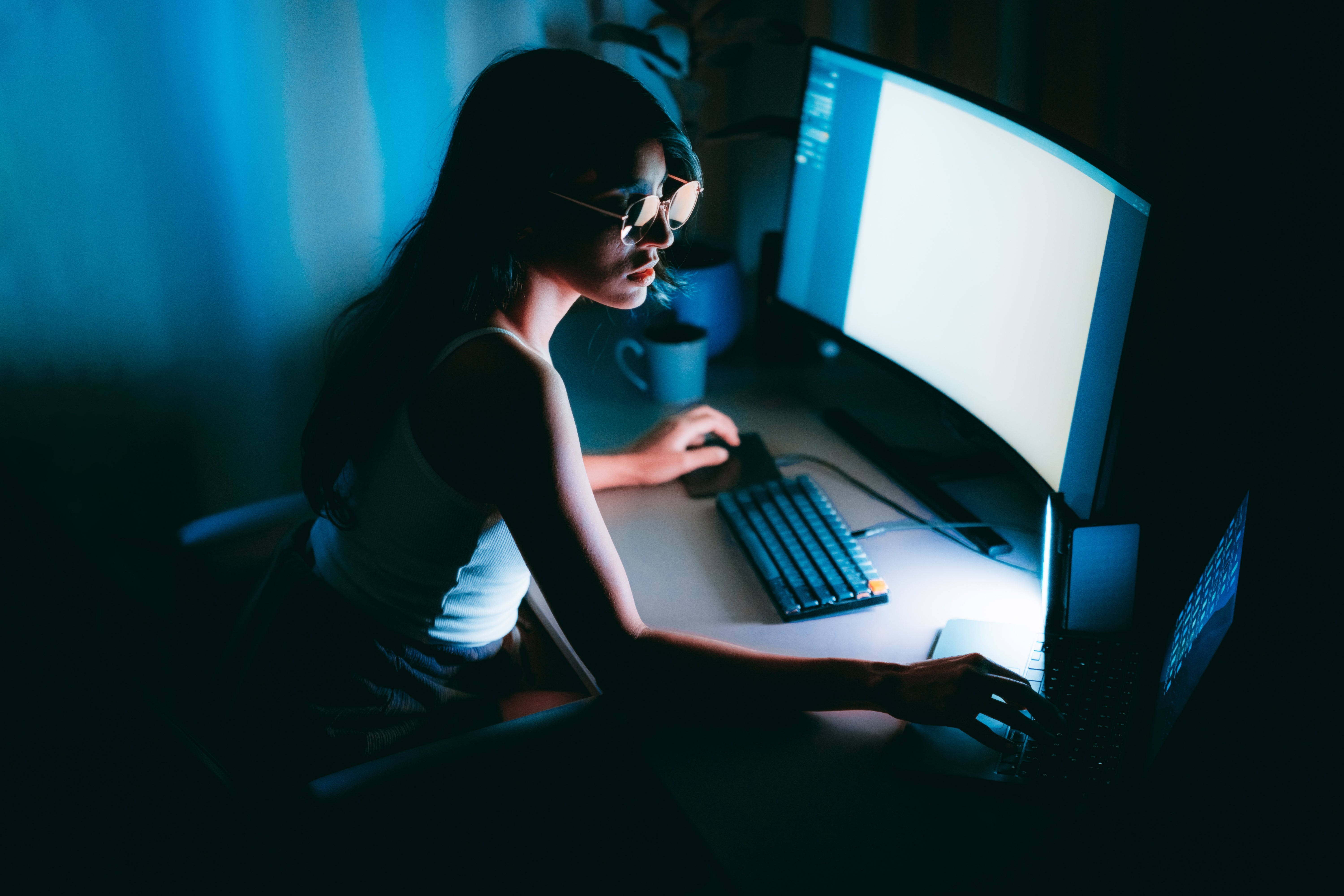 Person i briller som jobber ved et databord med to skjermer i svak belysning, fokusert på skjermene