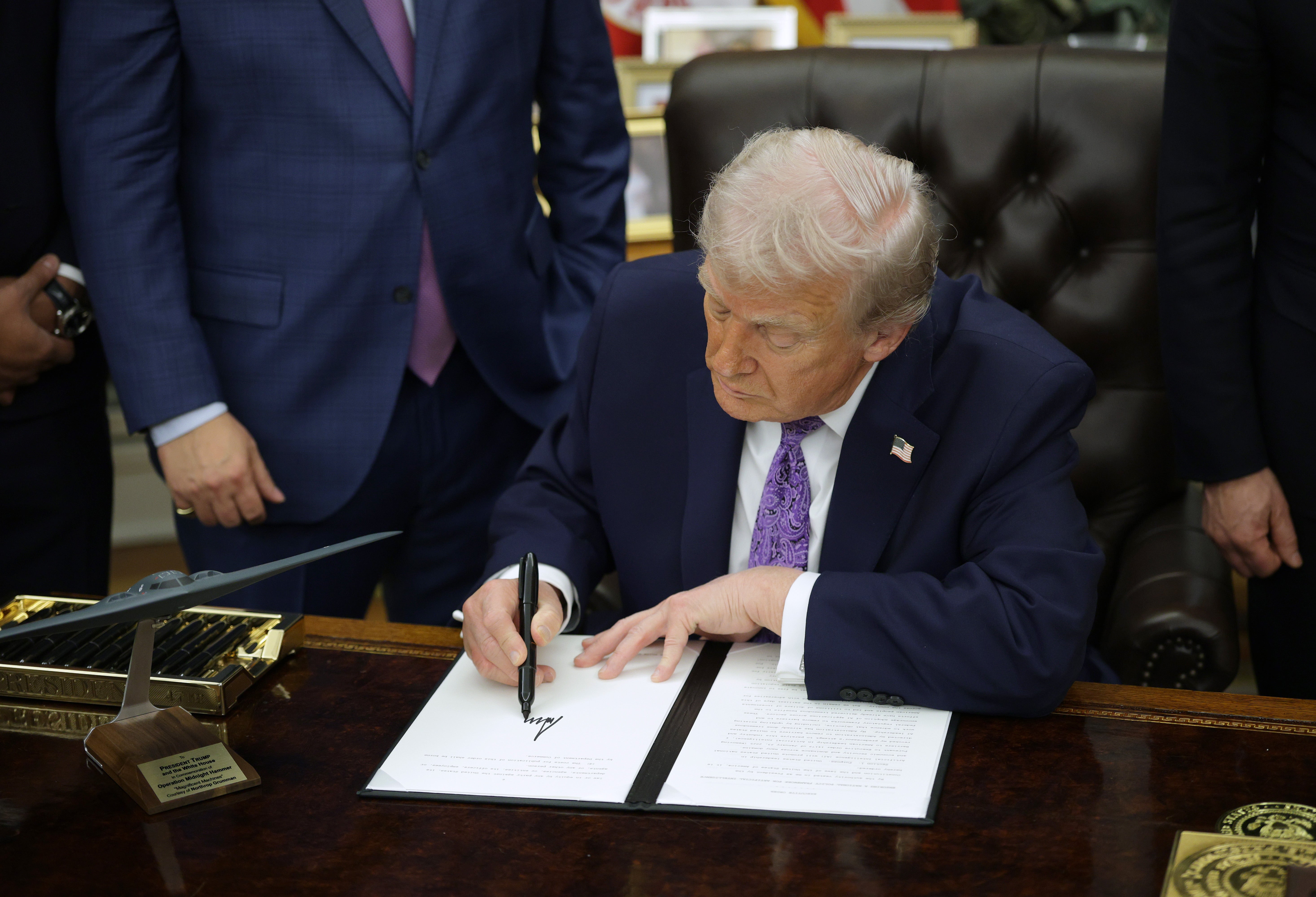 En person signerer et dokument ved et skrivebord i kontormiljø, omgitt av andre i dress, noe som antyder en offisiell eller politisk begivenhet