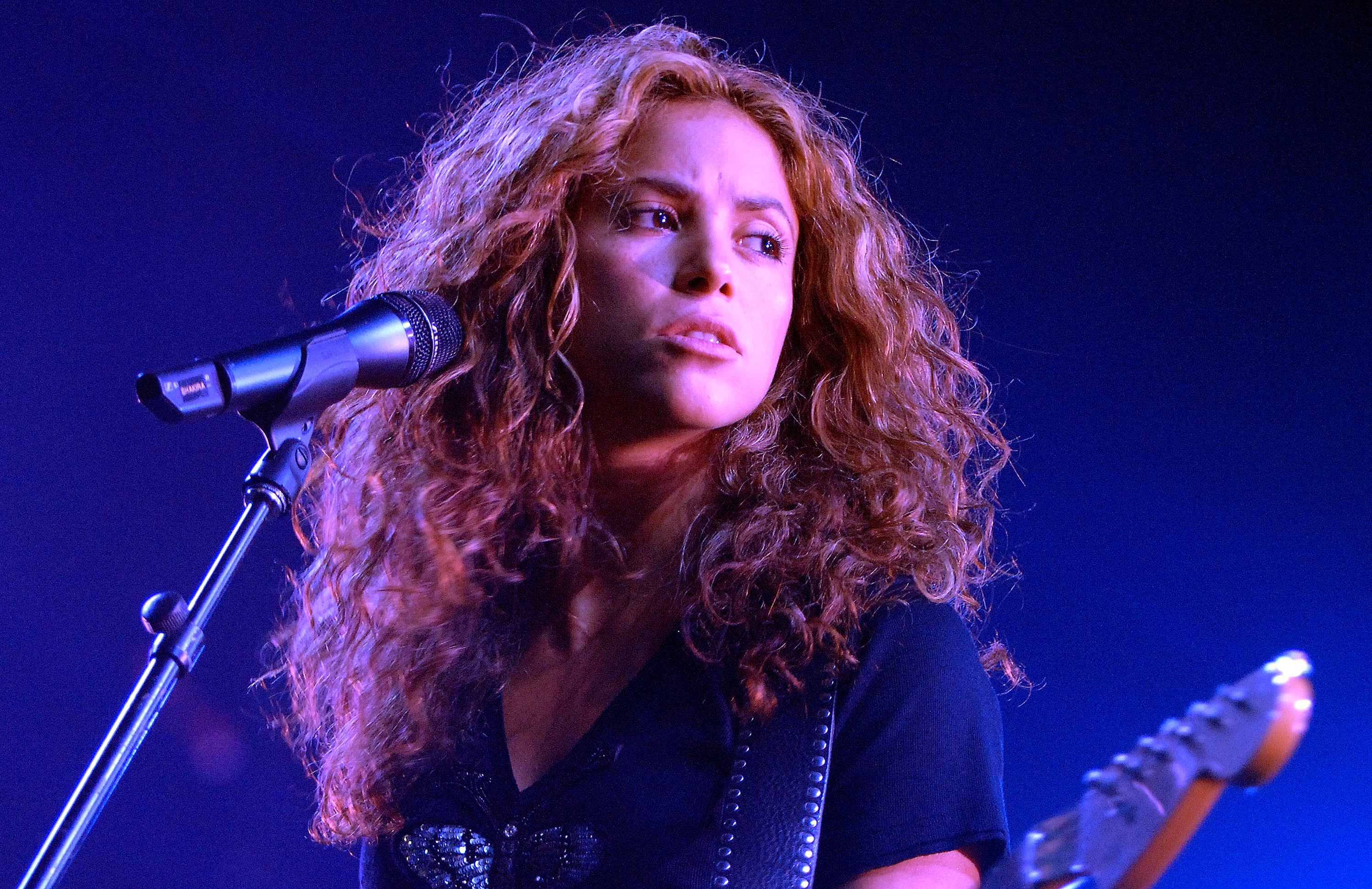 Singer performing on stage with a microphone, holding a guitar, wearing a black top, and looking to the side