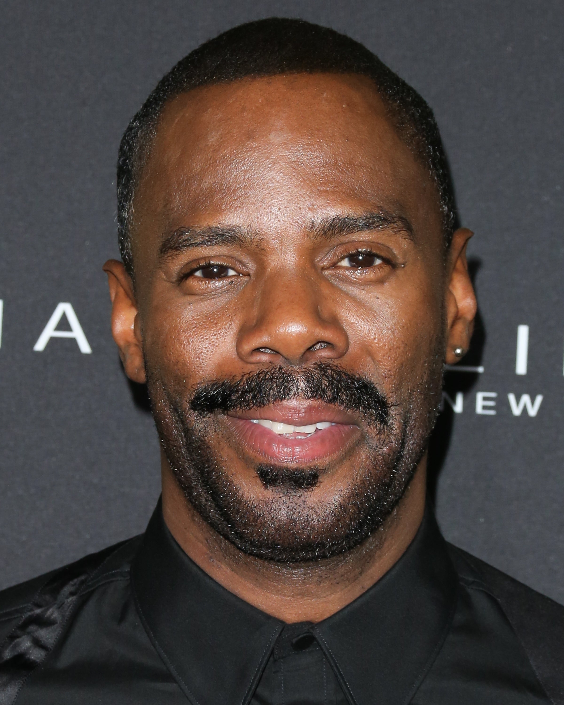 A person wearing a sleek black shirt poses on a red carpet in front of a branded backdrop, smiling for the camera