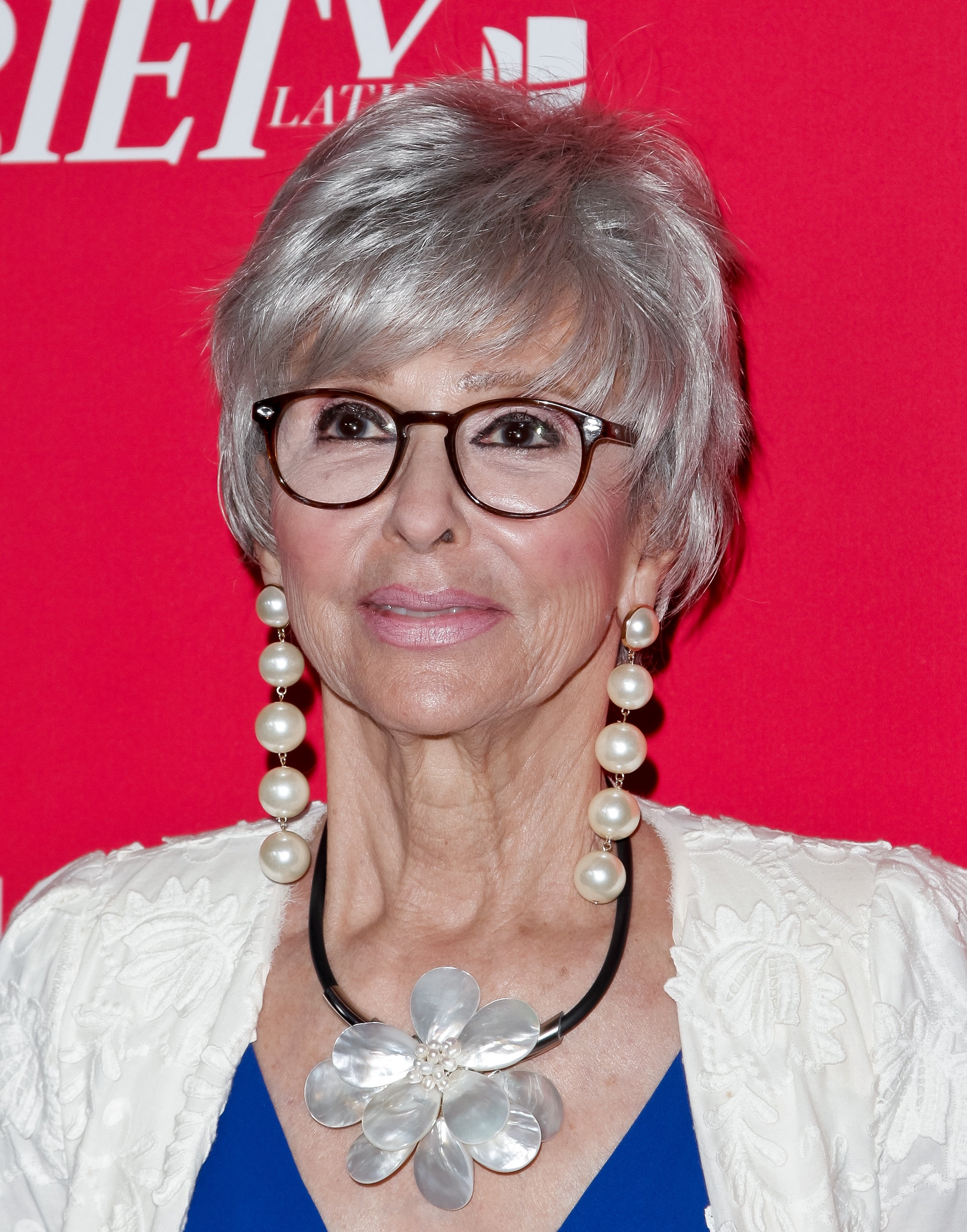 A woman with short hair and glasses is on a red carpet, wearing a floral necklace and pearl earrings with a patterned jacket over a dress