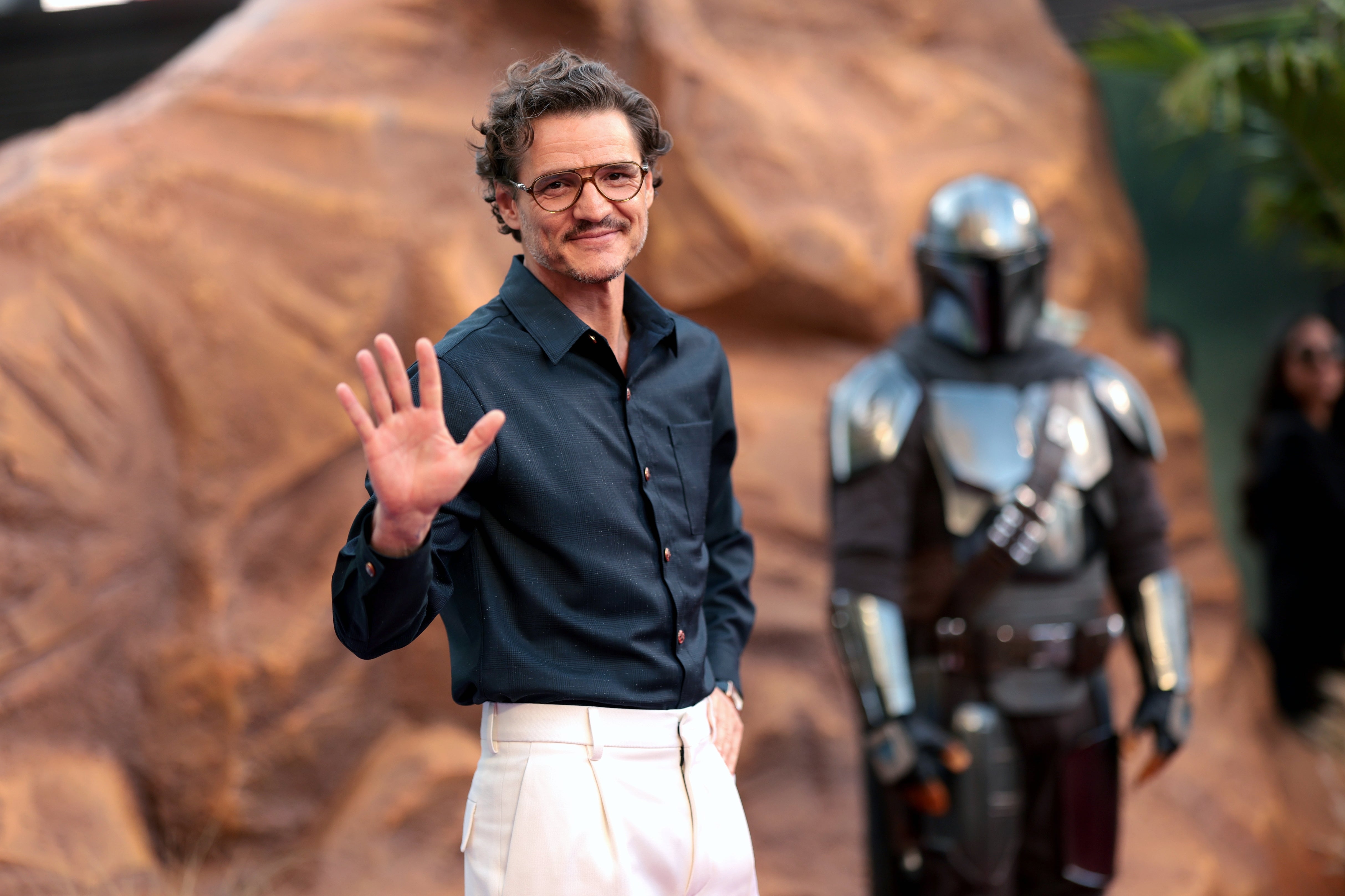 Person in a dark shirt and white pants waves on a red carpet. Another stands in armor nearby