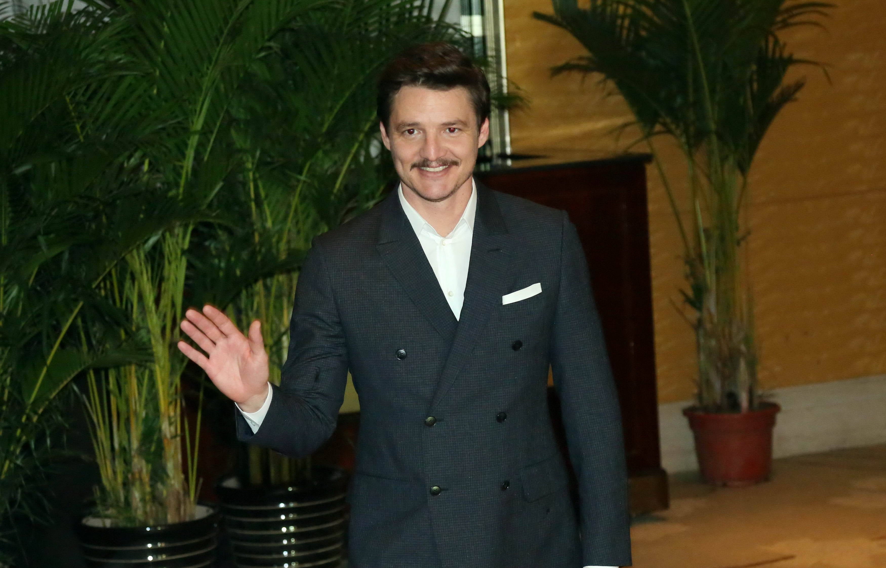 A person in a double-breasted suit waves while standing in front of large potted plants indoors