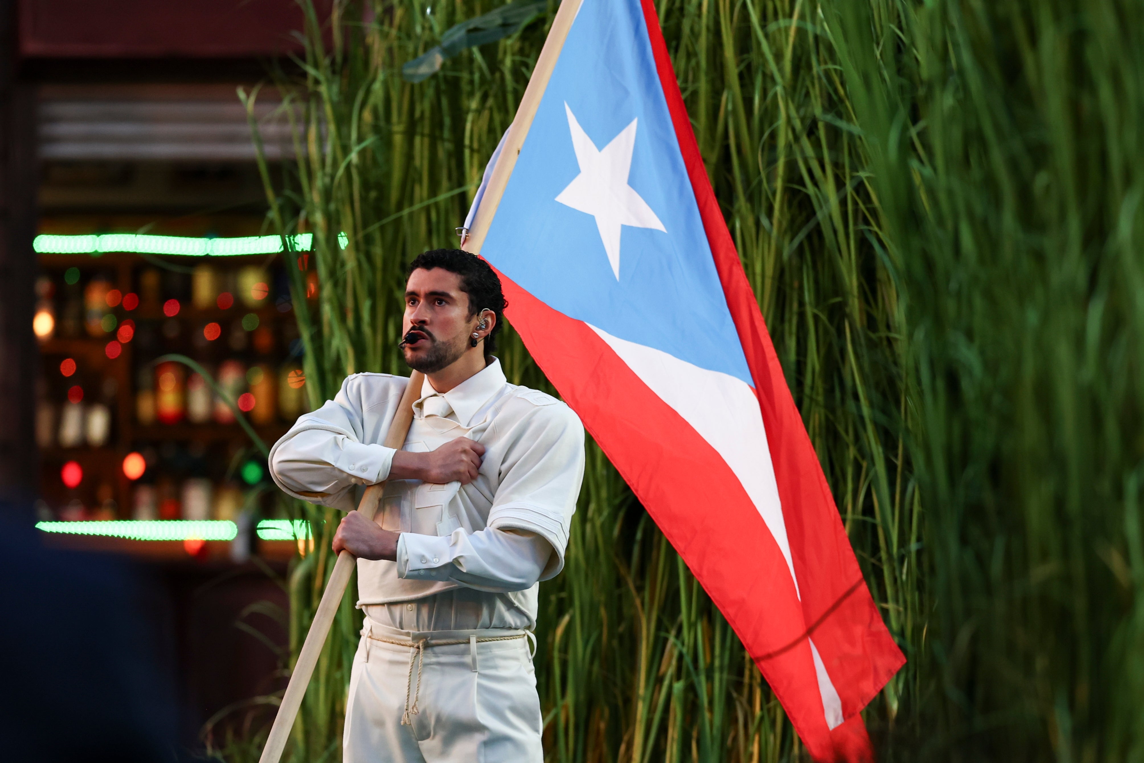A person in traditional attire holds a Puerto Rican flag against a backdrop of tall plants