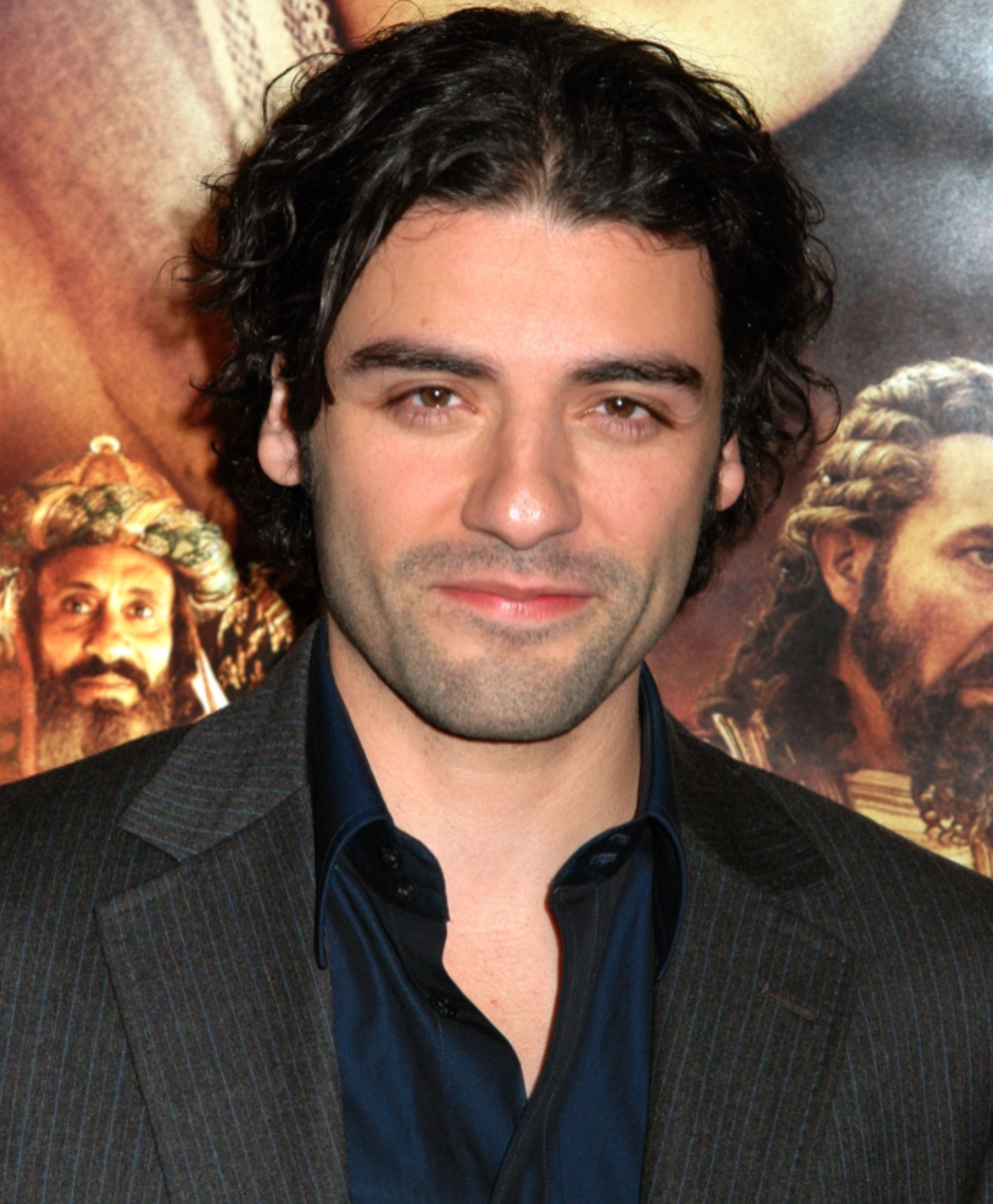 A person with curly hair in a suit stands in front of a movie poster backdrop
