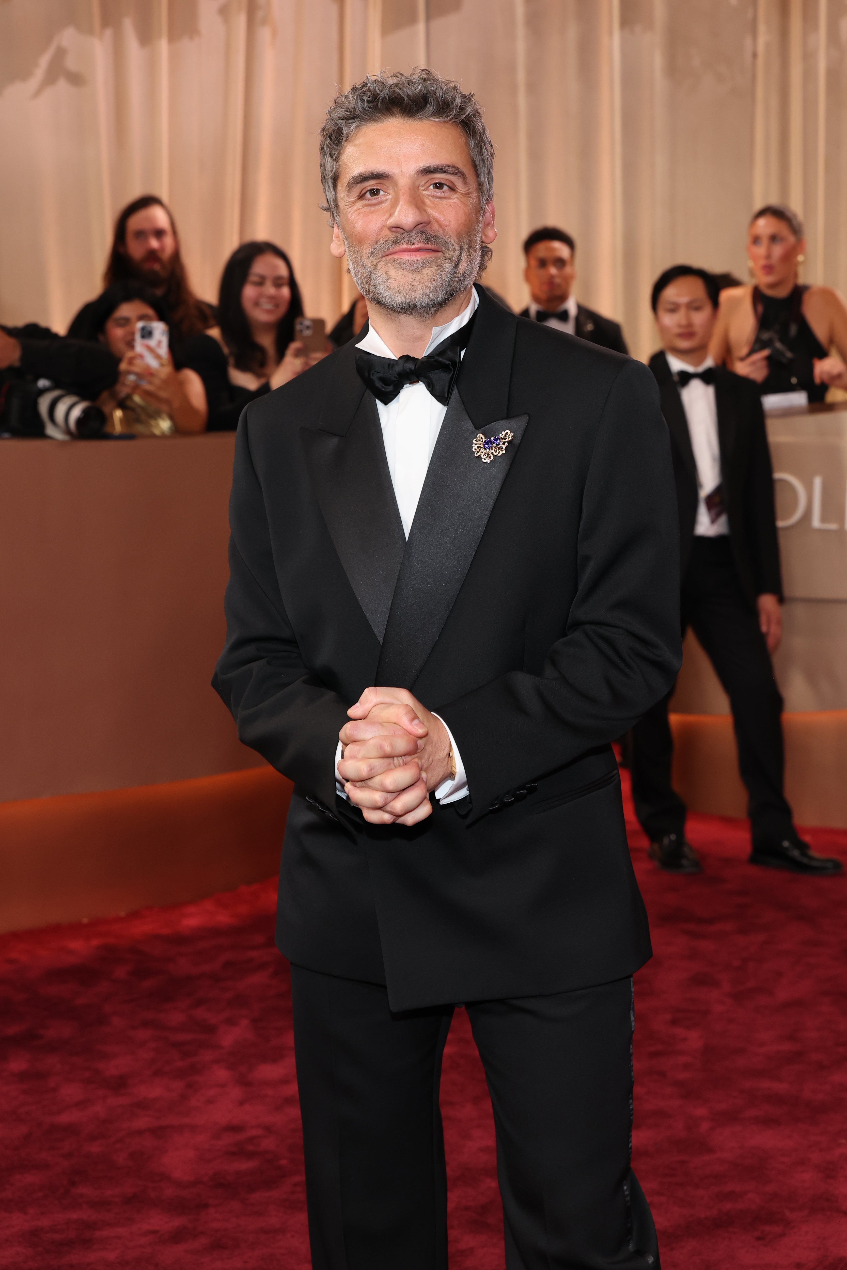 Person in a black tuxedo with a butterfly brooch on a red carpet, posing for photos