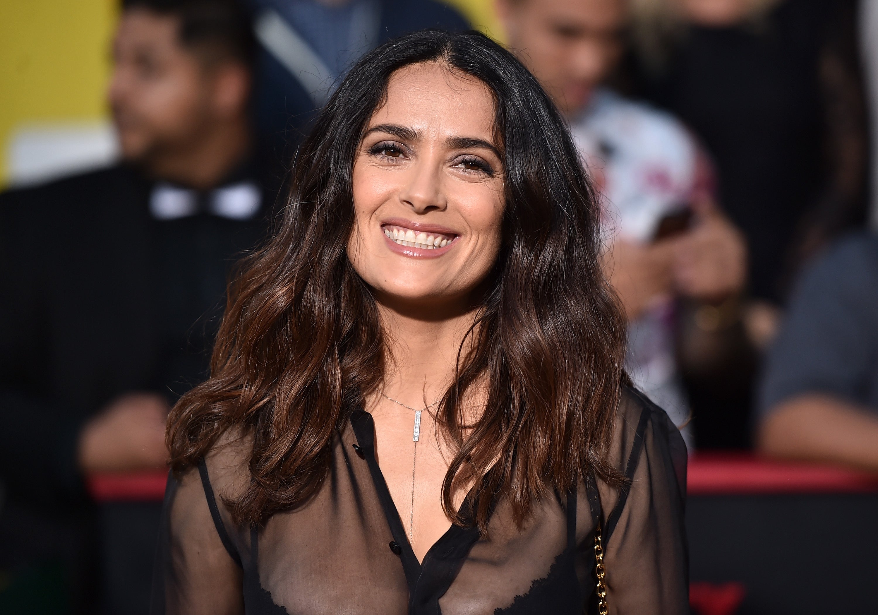A woman smiles on a red carpet, wearing a sheer black blouse with a necklace, surrounded by a blurred crowd