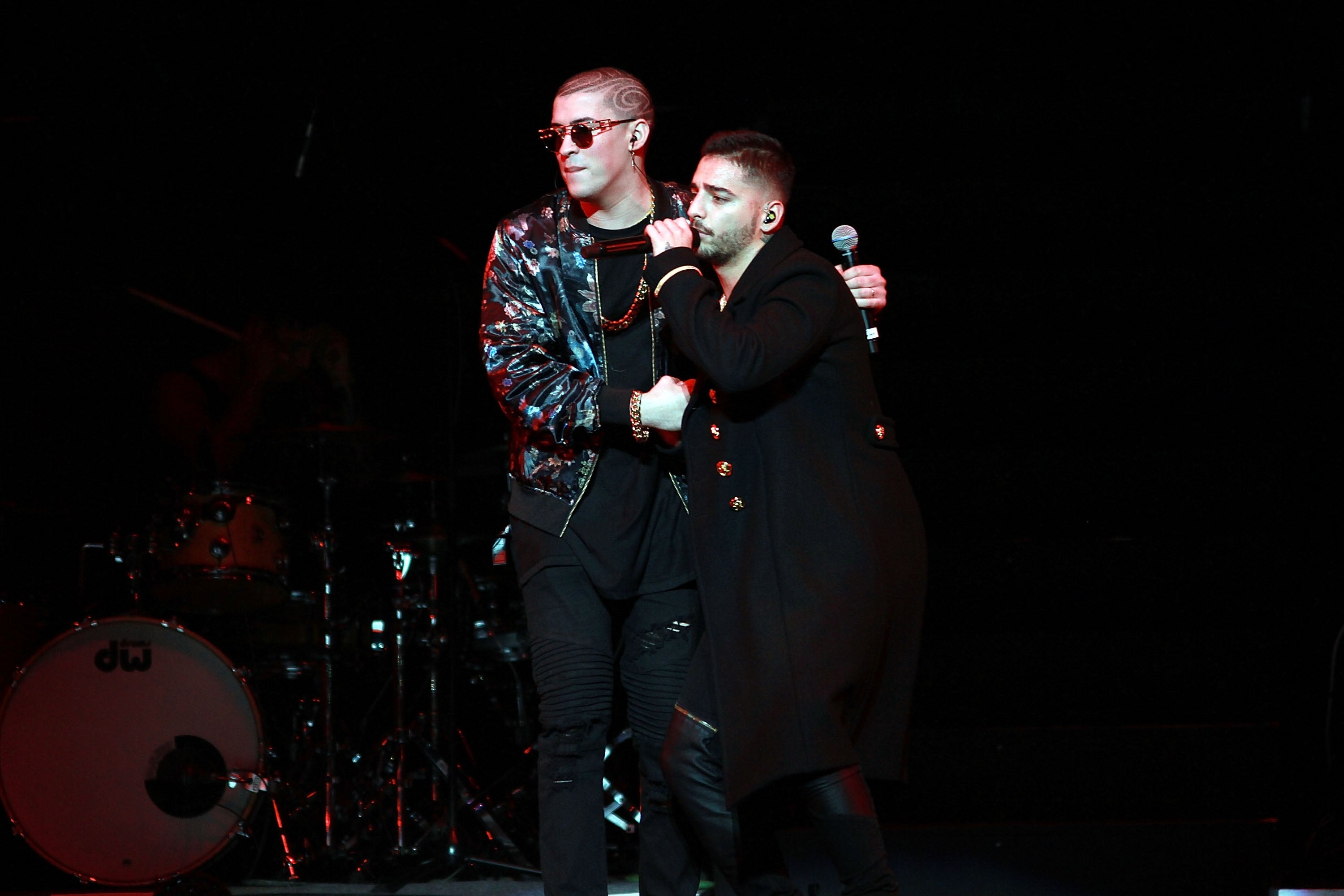 Two performers embrace on stage. One wears a shiny jacket and sunglasses; the other in a long coat holds a microphone