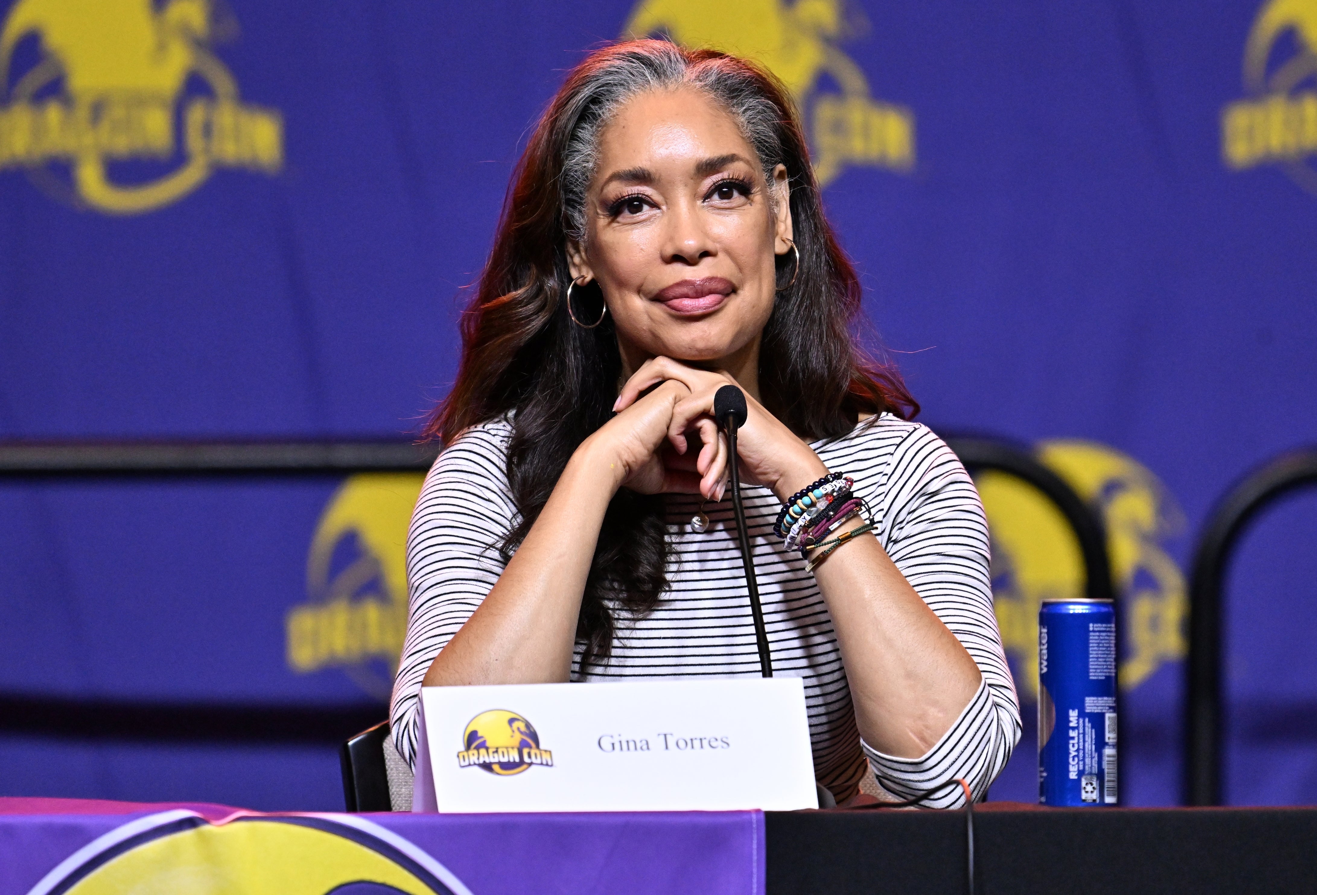 A person sits at a panel table with a microphone, wearing a striped top and bracelets, and appears attentive during a discussion